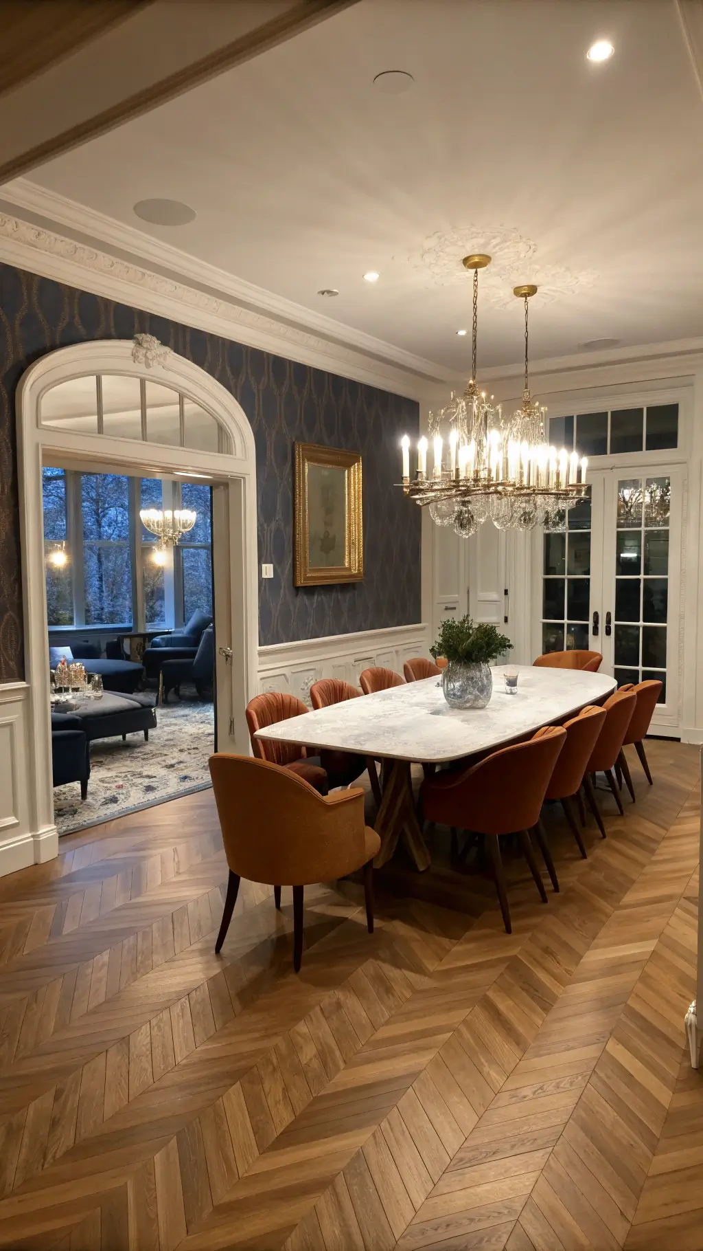 Expansive dining room at dusk with herringbone hardwood floors, a marble dining table, and restored 1950s cognac leather chairs. An antique gilt mirror reflects a modern chandelier. Navy grasscloth wallpaper with white wainscoting adds contrast. Candlelight and dimmed sconces create an intimate ambiance.