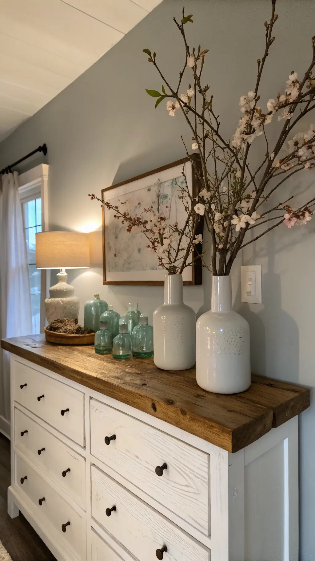 Master bedroom corner at blue hour with moody lighting. A reclaimed wood floating shelf holds vintage milk glass vases with cherry blossoms. Below, a whitewashed dresser with aged brass hardware is adorned with natural linen runners. Soft artificial fill light enhances the warm, intimate ambiance.