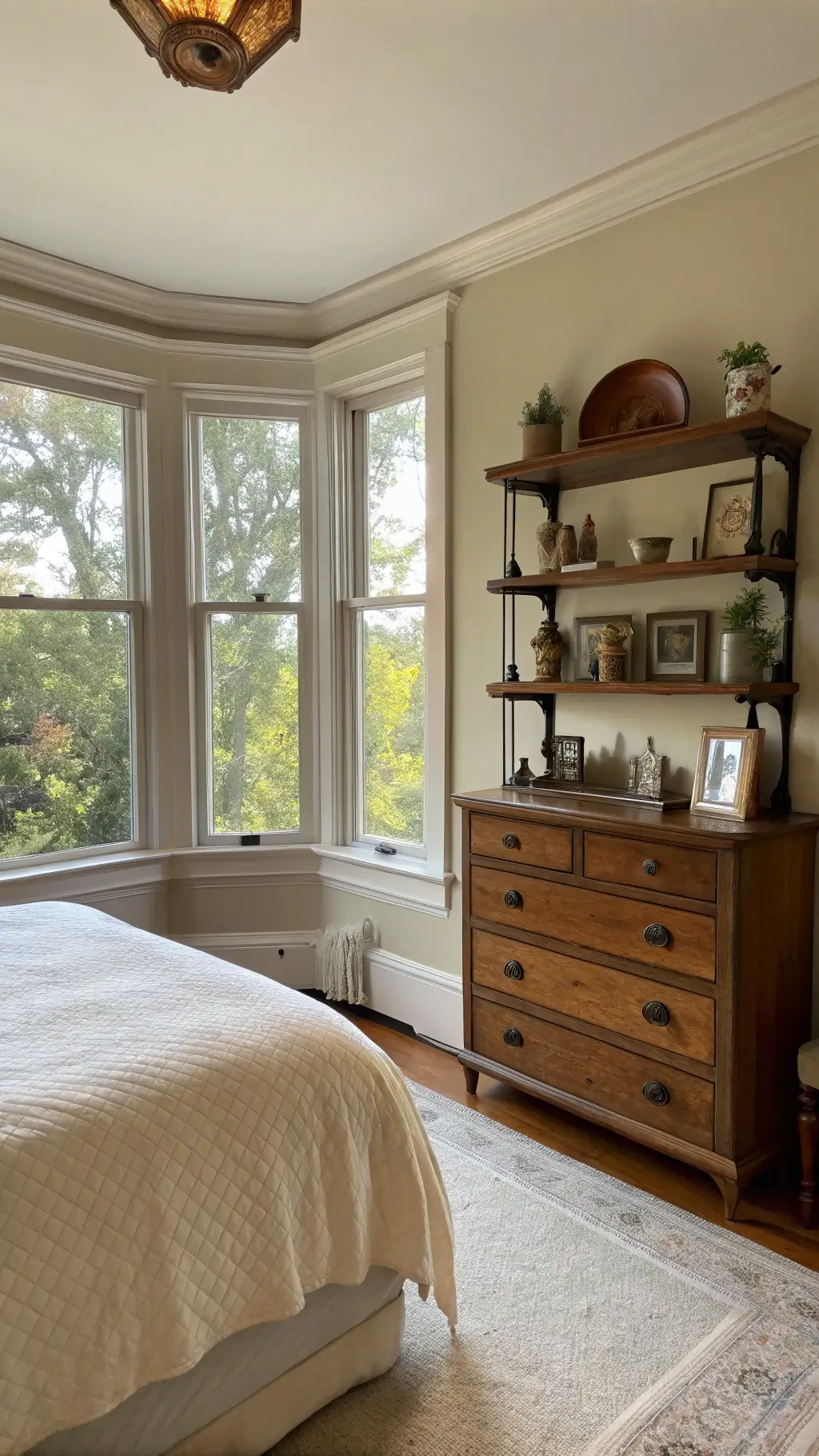 Traditional bedroom in late afternoon light with a prominent Eastlake period dresser, modern floating shelves displaying vintage objects, and soft cream bedding with subtle texture. Bay windows cast a gentle backlight, enhancing the serene and collected atmosphere.