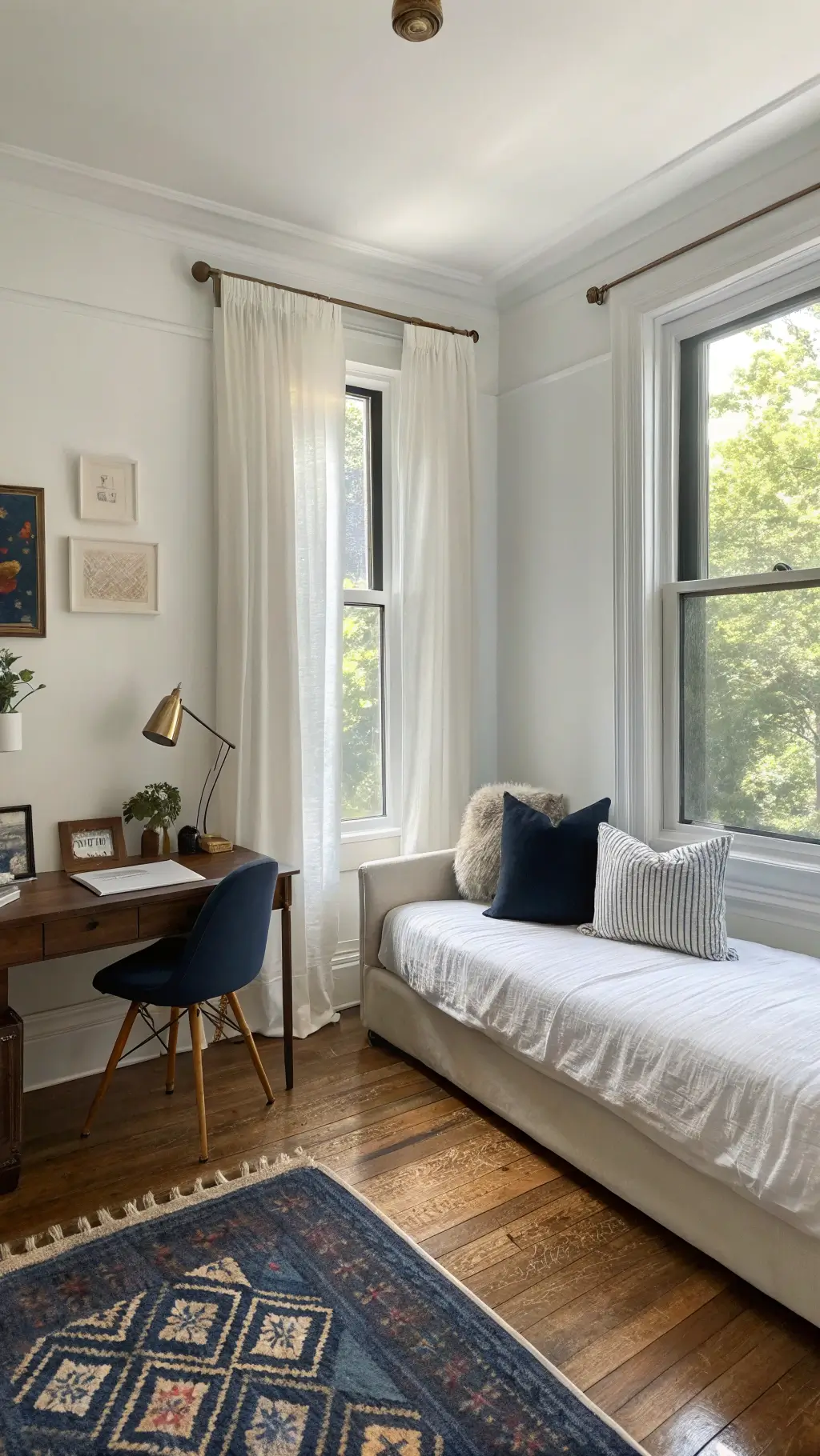 Guest room and home office hybrid with a modern daybed, navy accent pillows, a walnut desk by the window, and a vintage Persian rug on light oak flooring. Sunlight filters through sheer linen curtains, highlighting brass and glass details.