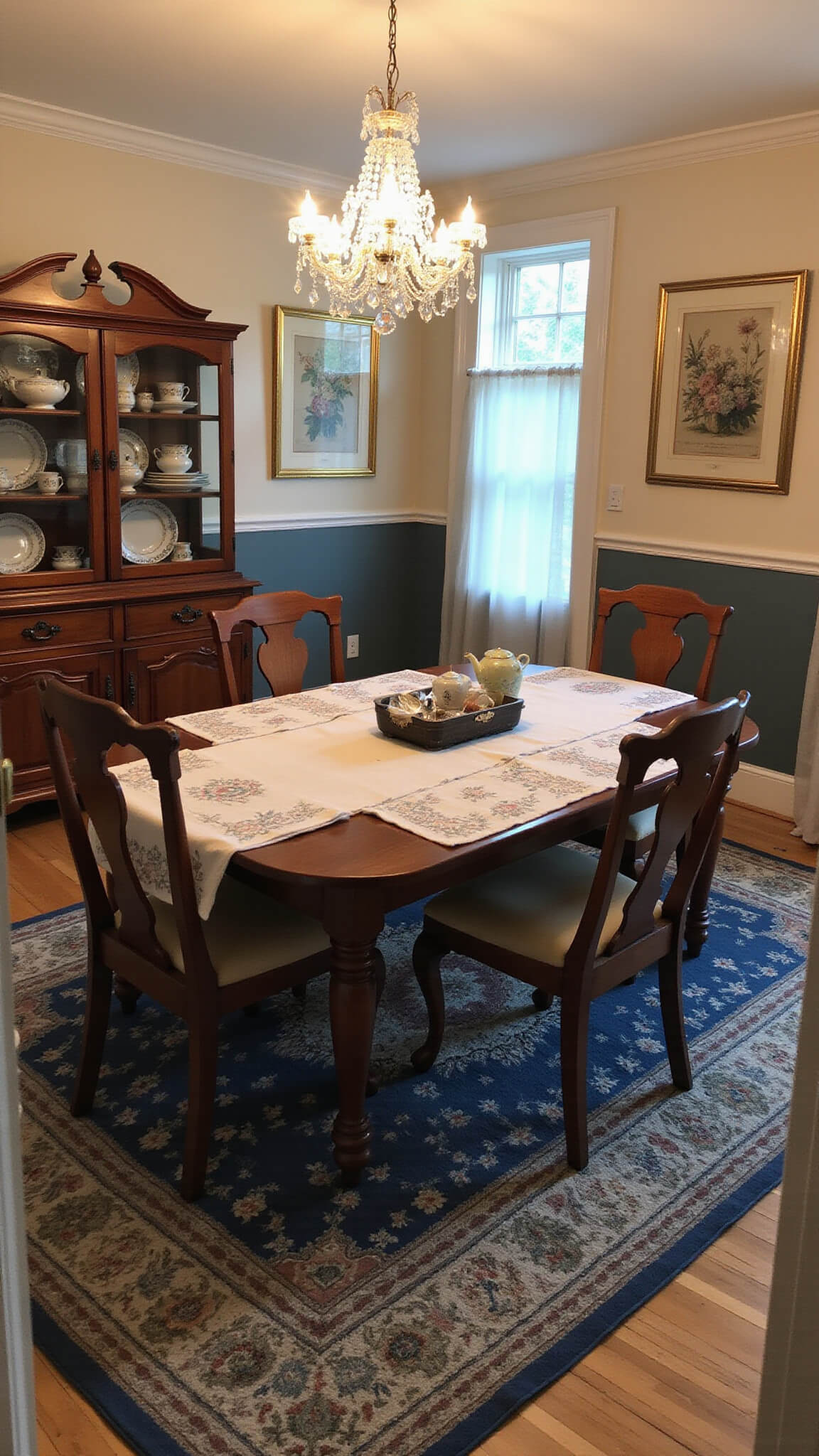Wide-angle view of a 15x18ft dining room at dusk with crystal chandelier illuminating mahogany table set with embroidered linens, vintage china cabinet, silverware box, botanical prints on walls, and braided rug in muted blues and browns.