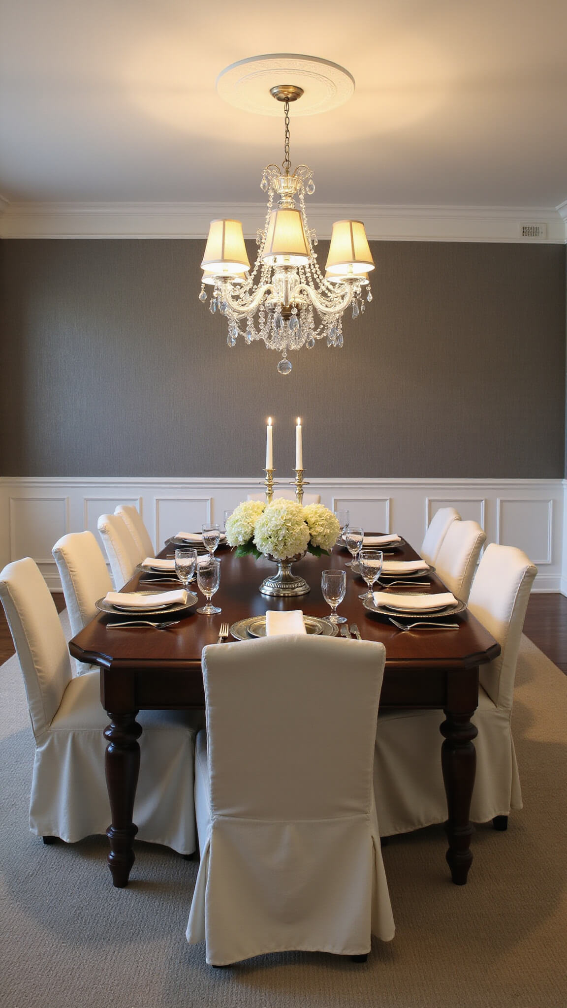 Elegant formal dining room at dusk with dark walnut table set for eight, cream linen slip-covered chairs, vintage silver candlesticks, white hydrangeas centerpiece, and crystal chandelier, featuring pewter grasscloth wallpaper and wainscoting.