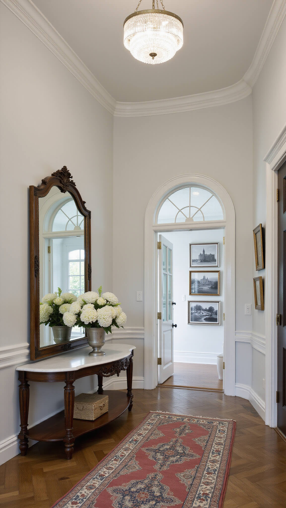Elegant foyer with circular layout featuring round marble table, fresh white flowers, vintage runner on herringbone floor, large mirror reflecting light from arched doorway, crystal ceiling fixture, and gallery wall of black and white photos.