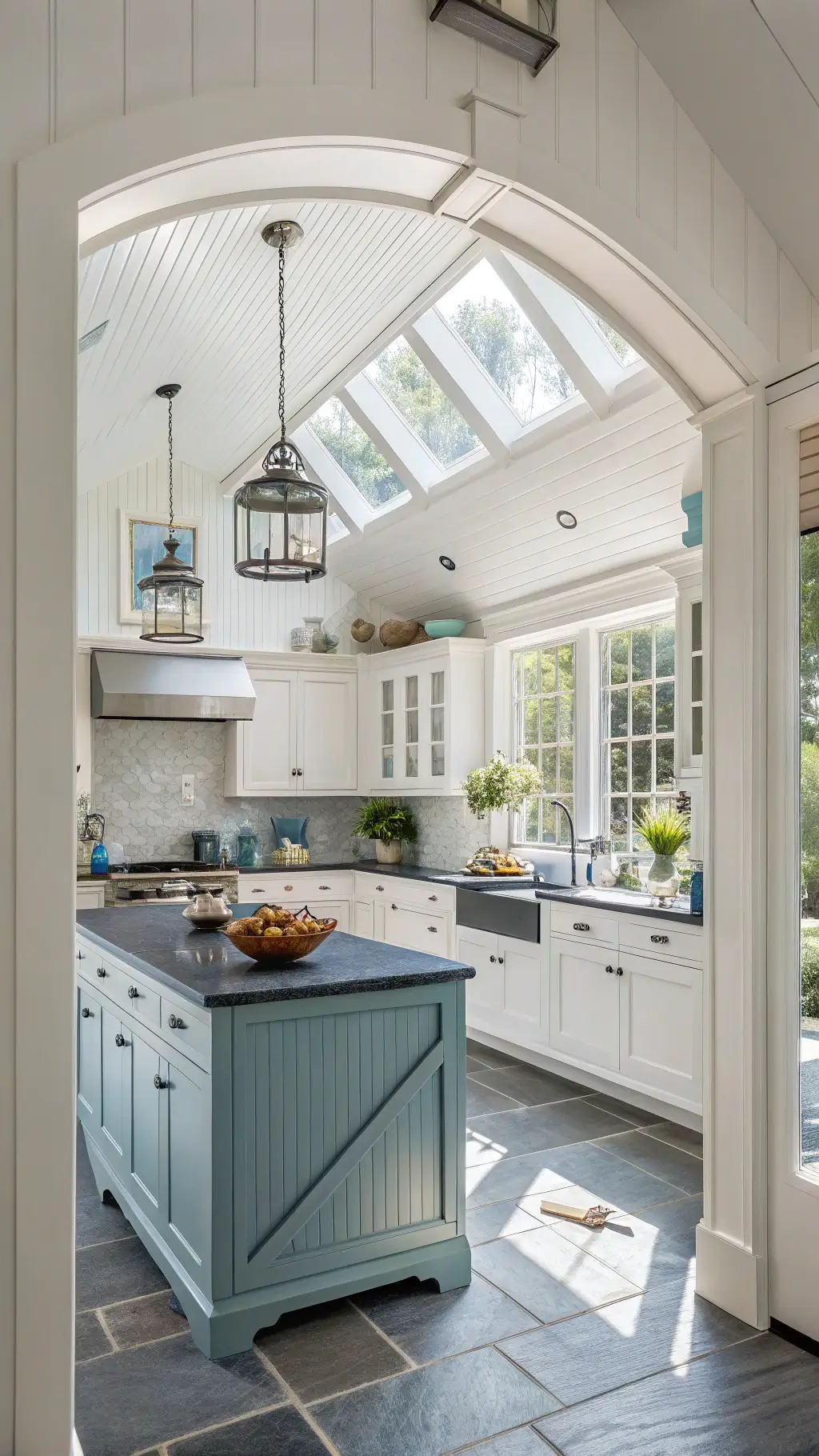 Bright coastal kitchen with vaulted ceiling, skylight, white beadboard cabinets, black granite countertops, blue-grey island, and rattan pendant lights.