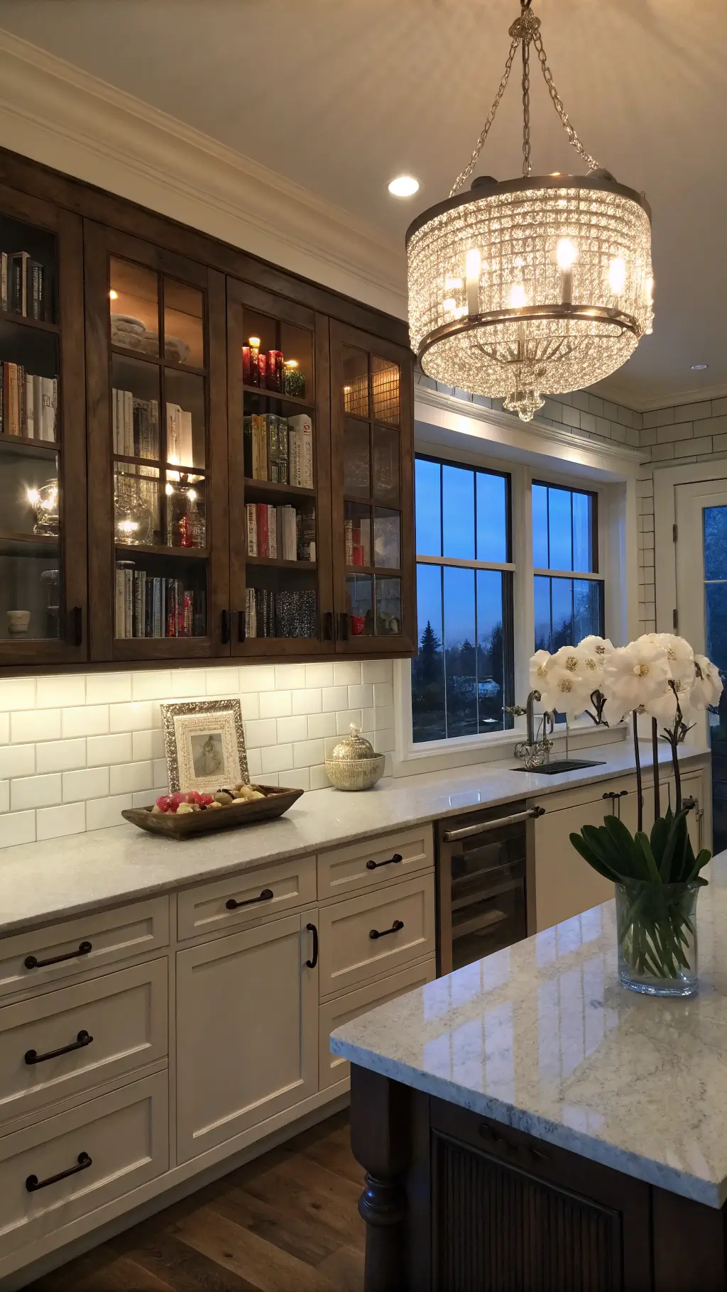 Transitional kitchen at dusk with whitewashed oak cabinets, dark walnut island, crystal chandelier, white subway tile, leather cookbooks, orchids, and moody blue hour lighting.