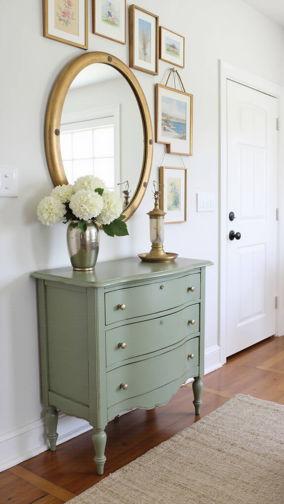 Vintage sage green dresser with round gilt mirror, white hydrangeas in a mercury glass vase, and gallery wall of watercolor art in soft morning light.