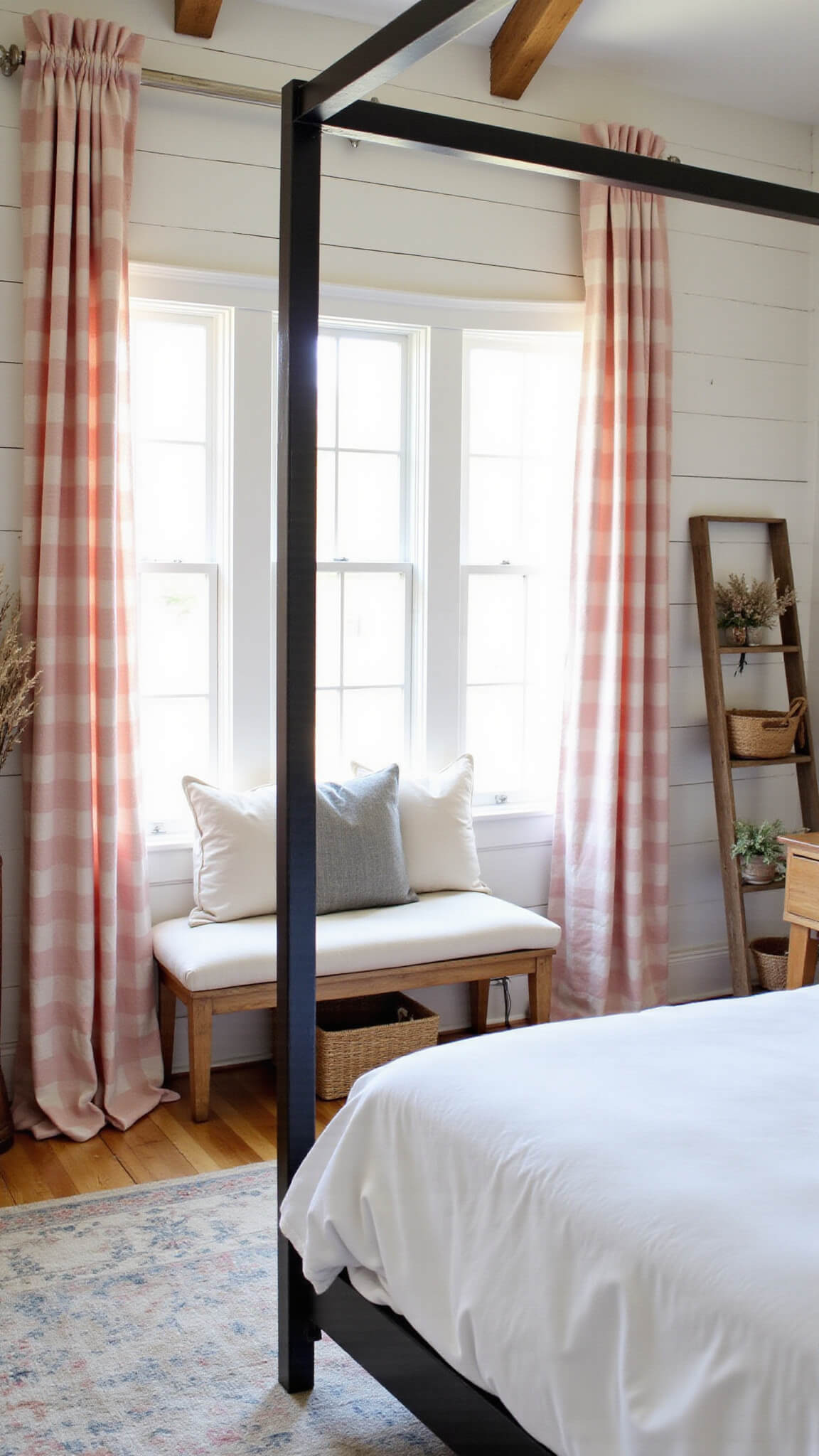 Modern farmhouse bedroom with blush pink buffalo check curtains, window seat, white shiplap walls, exposed beams, iron bed with white linens, vintage ladder, woven baskets, and dried florals.