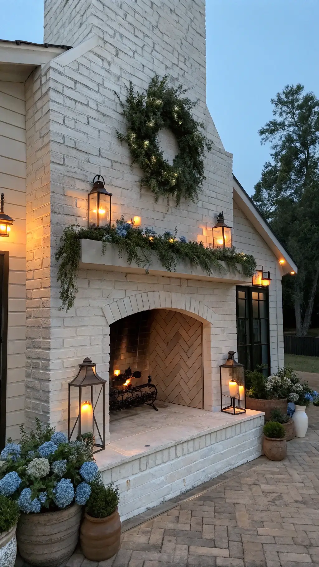 Modern farmhouse great room with whitewashed brick fireplace at dusk, featuring wrought iron lanterns, boxwood wreath, eucalyptus and thistle garland, terra cotta pots with spring bulbs, and warm Edison lighting.