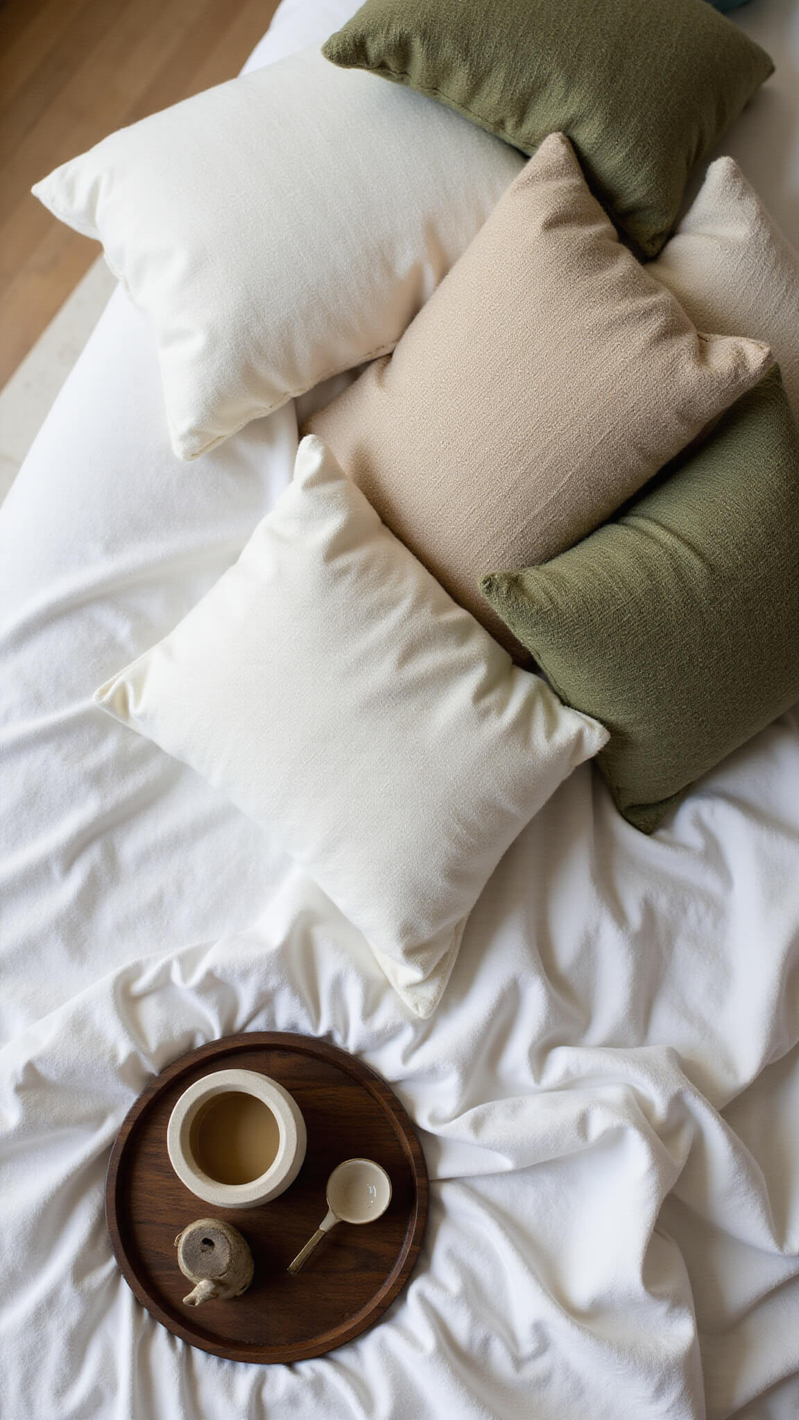 Overhead view of Japandi-style bedding with layered neutral linens, olive green wool pillows, and a dark wood tray holding a ceramic tea set in natural afternoon light.