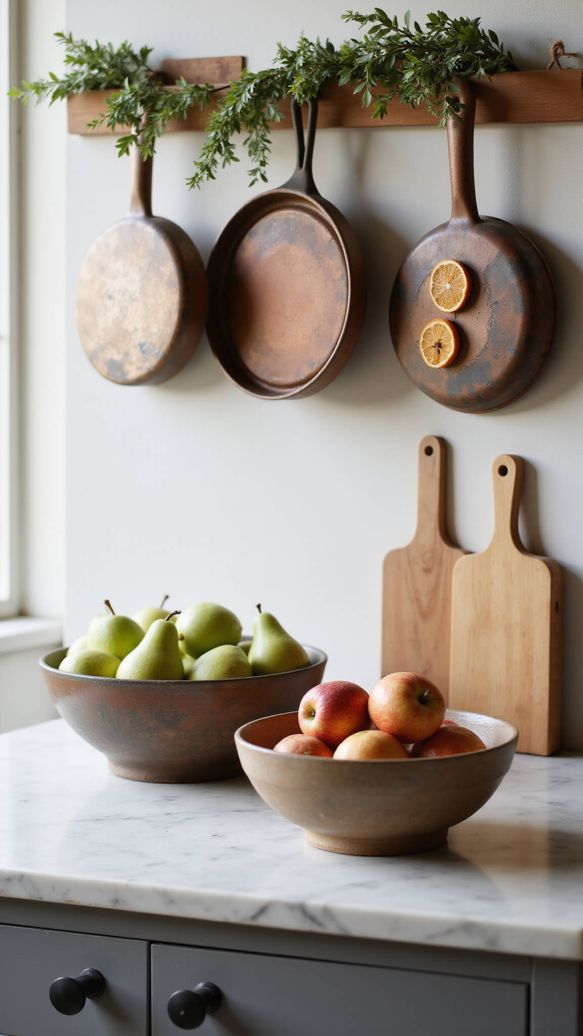 Marble kitchen island styled with ceramic bowls of apples and pears, copper cookware above, eucalyptus garland with dried oranges, and wooden cutting boards, softly lit from the side.