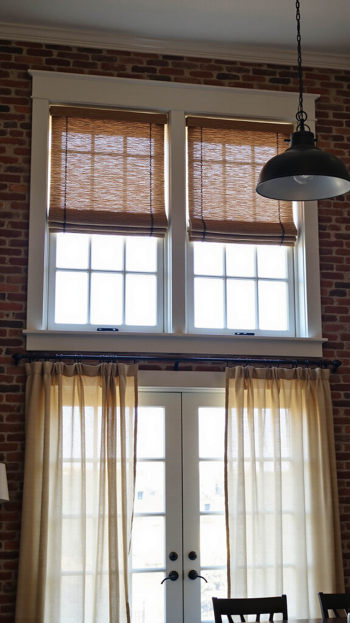Low-angle view of a double-height kitchen with 12ft windows, natural muslin café curtains below, woven wood blinds above, exposed brick walls, industrial pendant lights, black metal curtain rods, and dramatic shadow play in an industrial farmhouse style.