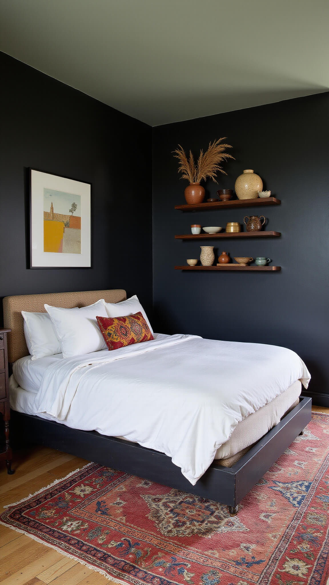 Corner king bed in 16x16ft room with matte black walls, sage green ceiling, layered kilim rugs over sisal floor, pottery on floating shelves, and pampas grass, photographed during blue hour from above.