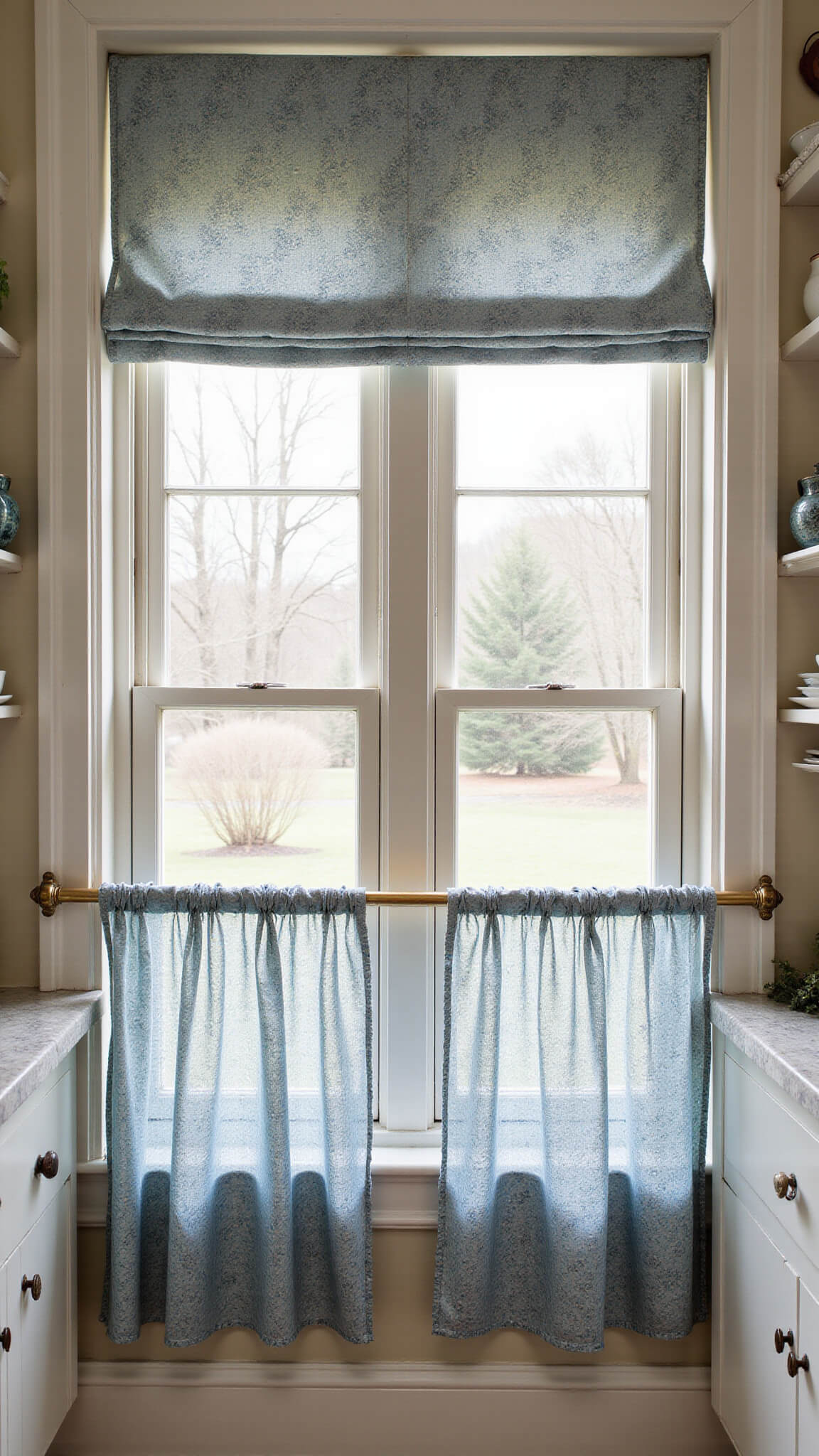 Galley kitchen with twin windows featuring mismatched floral and check curtains in muted blues and creams, aged brass rods with glass finials, vintage pottery on floating shelves, and soft afternoon light.