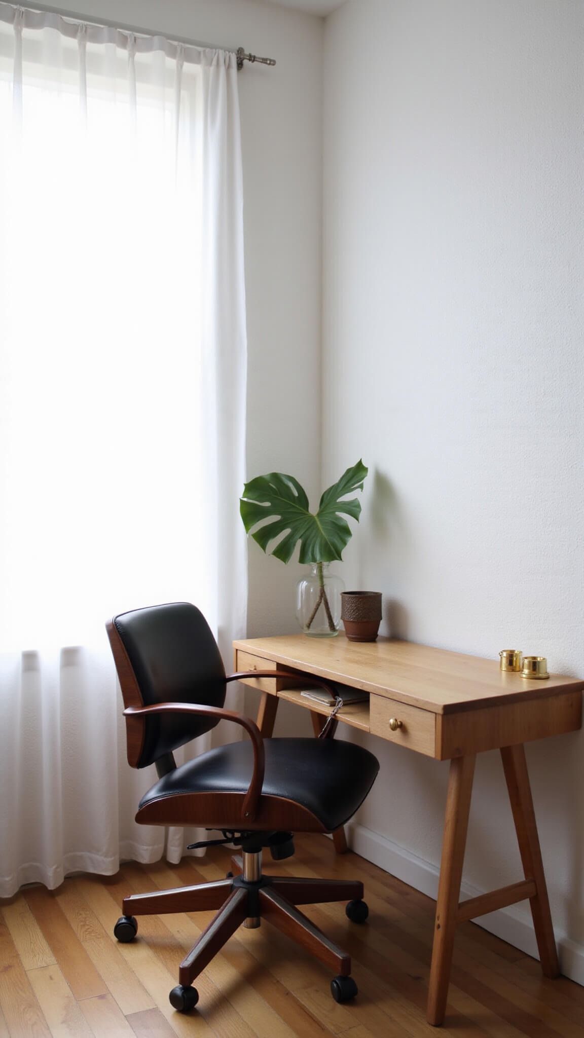 Low-angle view of a minimalist home office with soft morning light, featuring a black leather and walnut Japandi chair, oak desk with brass accents, and a potted monstera plant against a textured white wall.