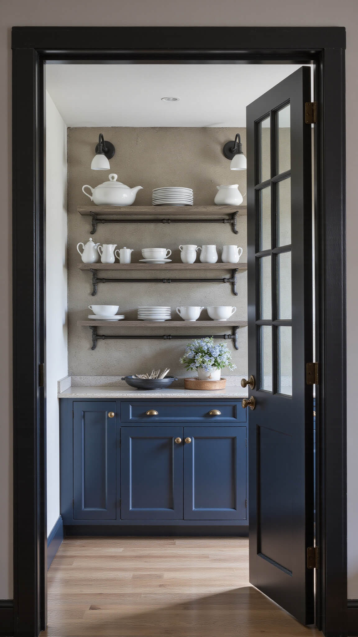 Moody butler's pantry with navy blue glass-front cabinets, white ceramics, vintage silverware, industrial pipe shelves, and antique mirror backsplash reflecting soft sconce lighting.