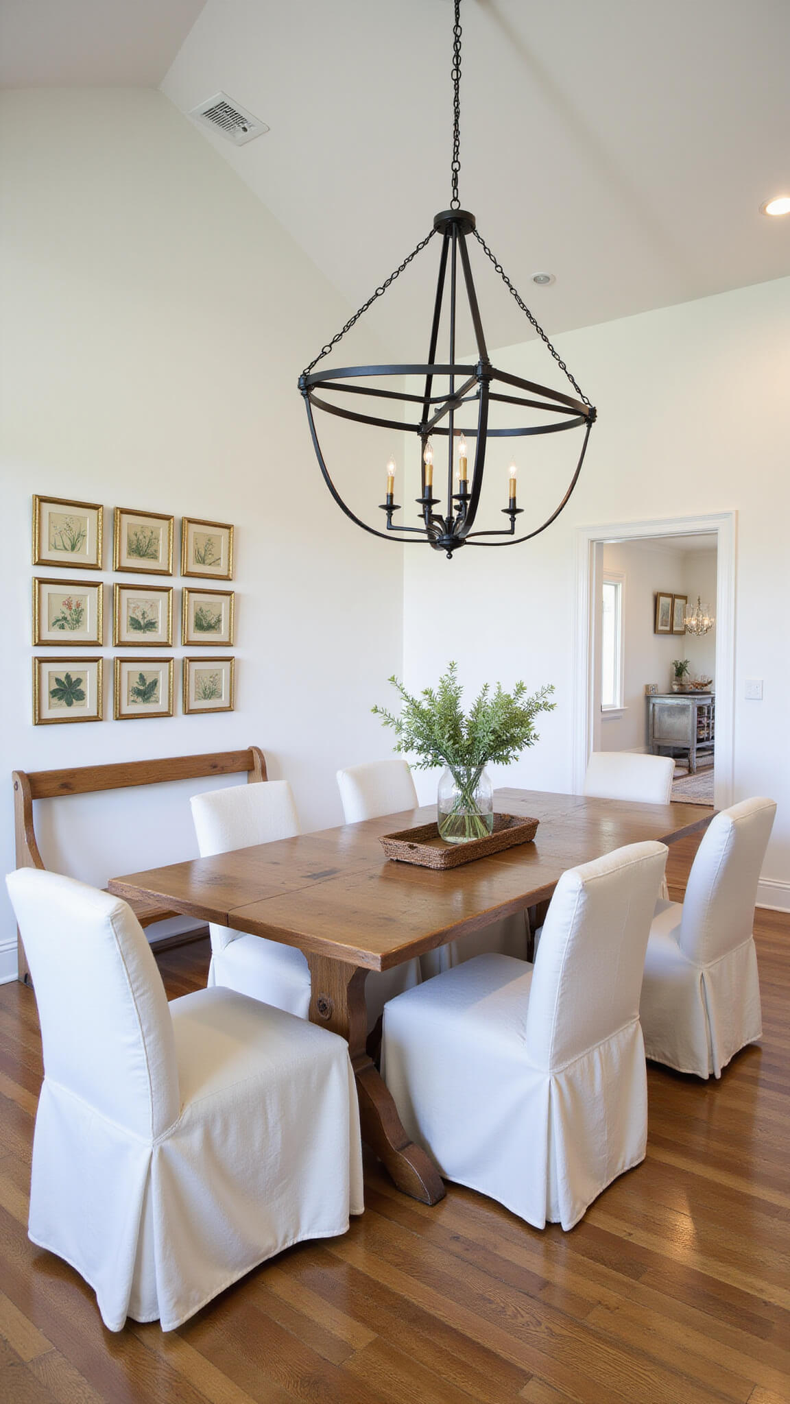 Modern farmhouse kitchen dining area with cathedral ceiling, featuring a wood table, slipcovered chairs, vintage church pew, oversized iron chandelier, gallery wall of botanical prints in brass frames, and natural oak accents.