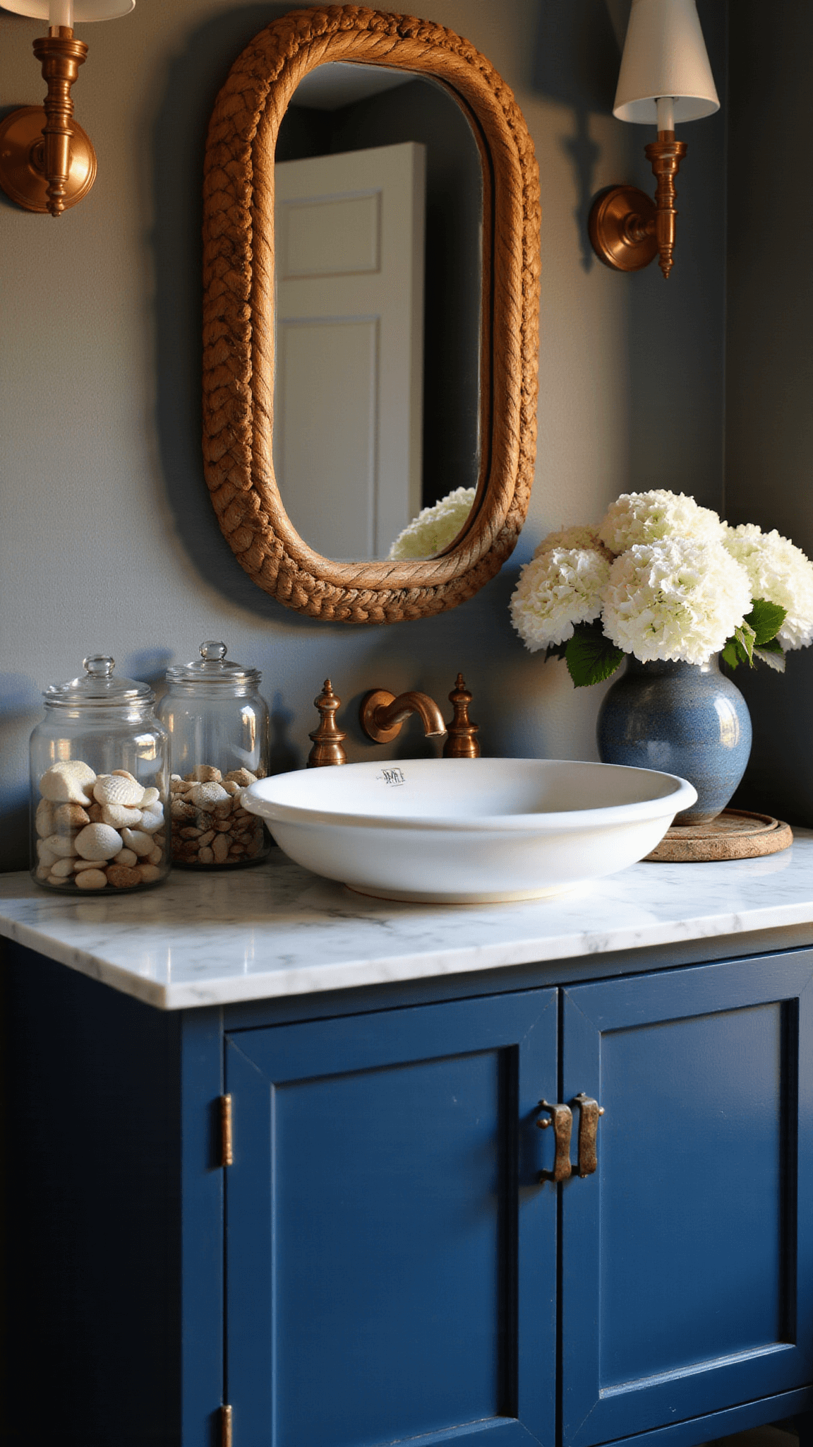 Moody evening bathroom with navy vanity, marble top, vintage apothecary jars, rattan mirror, and white hydrangeas in blue vase.