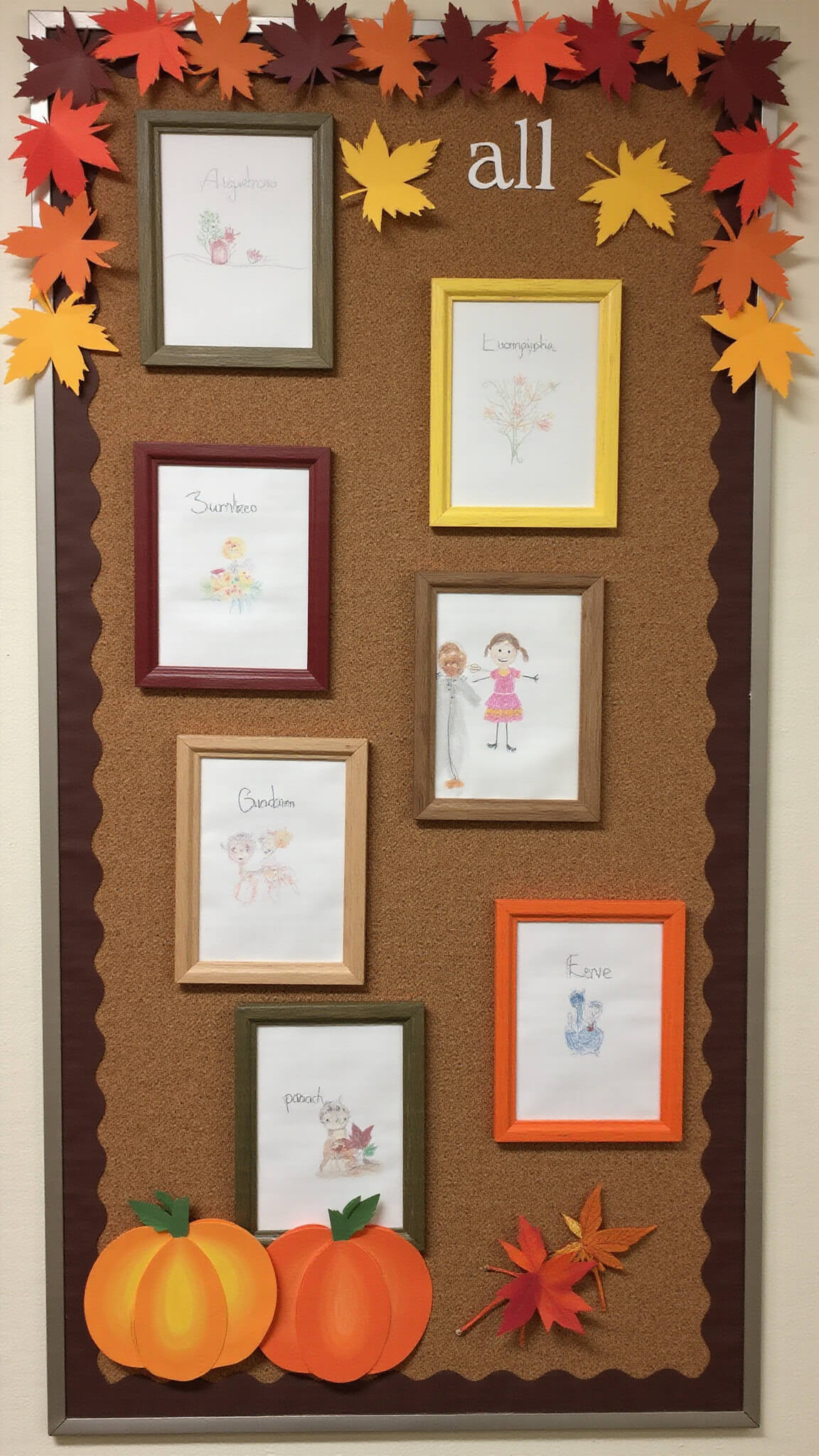 Straight-on view of an 8'x12' classroom bulletin board with brown cork background displaying student artwork in harvest-colored wooden frames, bordered by 3D paper maple leaves in fall shades and pumpkin-shaped name tags.
