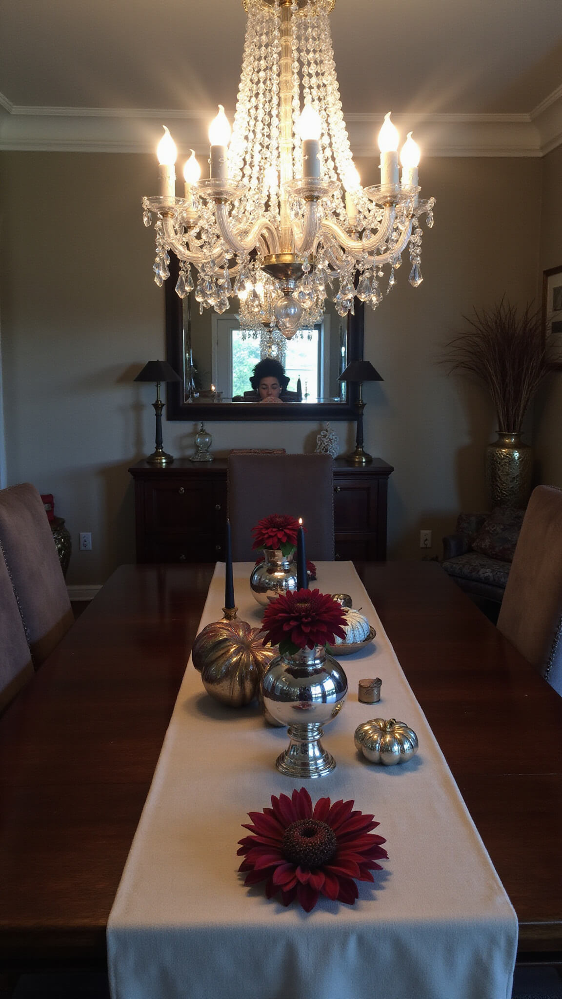 Moody formal dining room with crystal chandelier, dark wood table, velvet chairs, and autumnal centerpiece of mercury glass pumpkins, burgundy dahlias, and black taper candles.