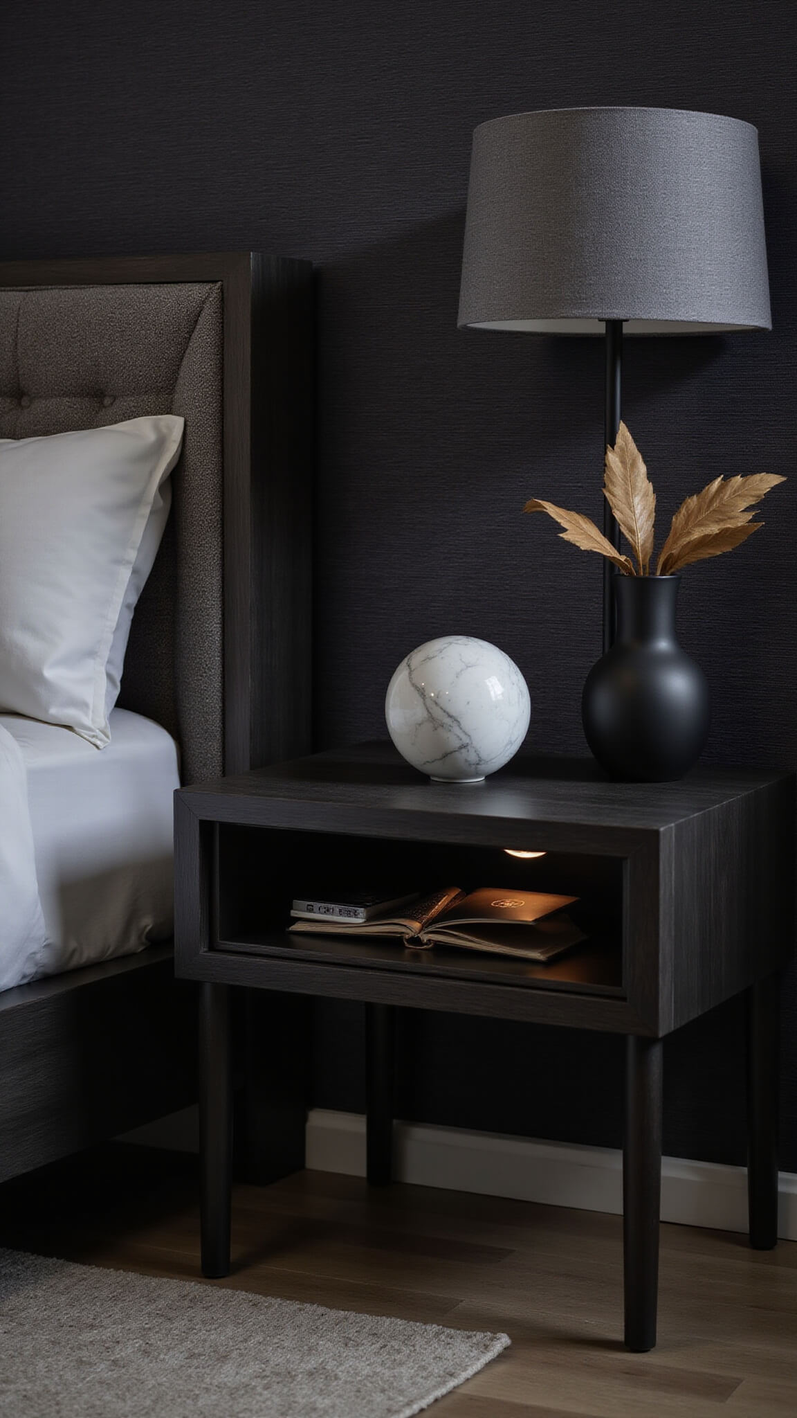 Moody evening bedroom corner with black oak nightstand, uplighting, and minimalist décor against charcoal grasscloth wallpaper.