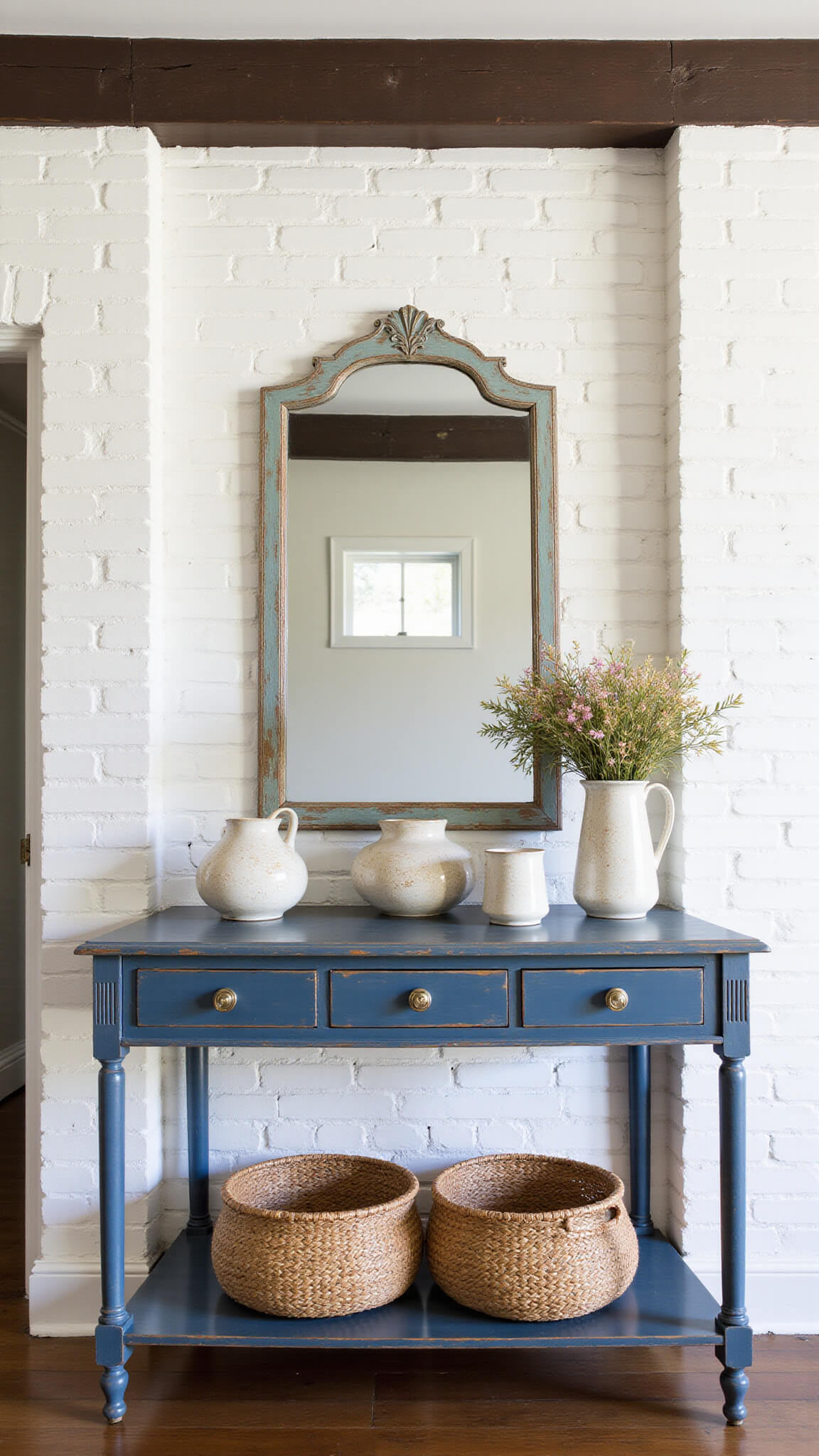 Welcoming entryway with whitewashed brick walls, dark wood beam, distressed blue console table with wildflowers, woven baskets below, and weathered copper mirror reflecting light.