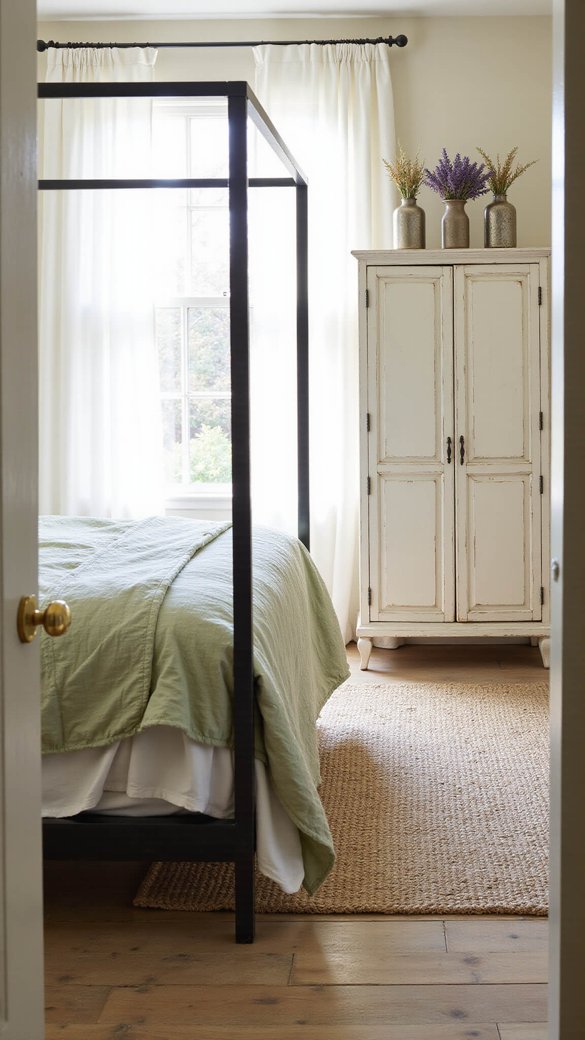 Sunlit cottagecore bedroom with vintage iron canopy bed, layered linens, and soft morning light streaming through white curtains.