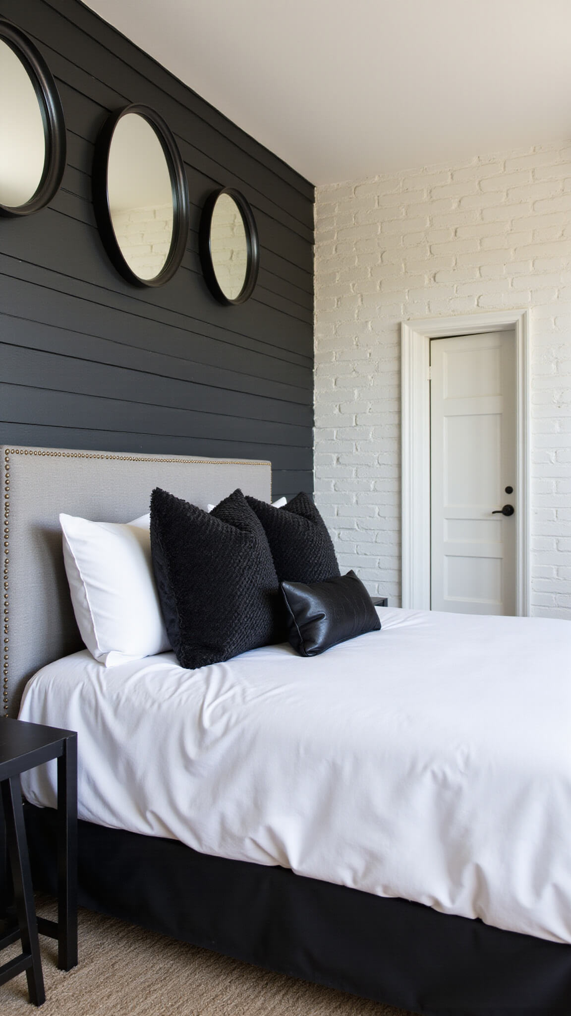 Industrial chic bedroom with black shiplap accent wall, white brick, gray upholstered bed, layered black pillows, and asymmetrical black-framed mirrors.