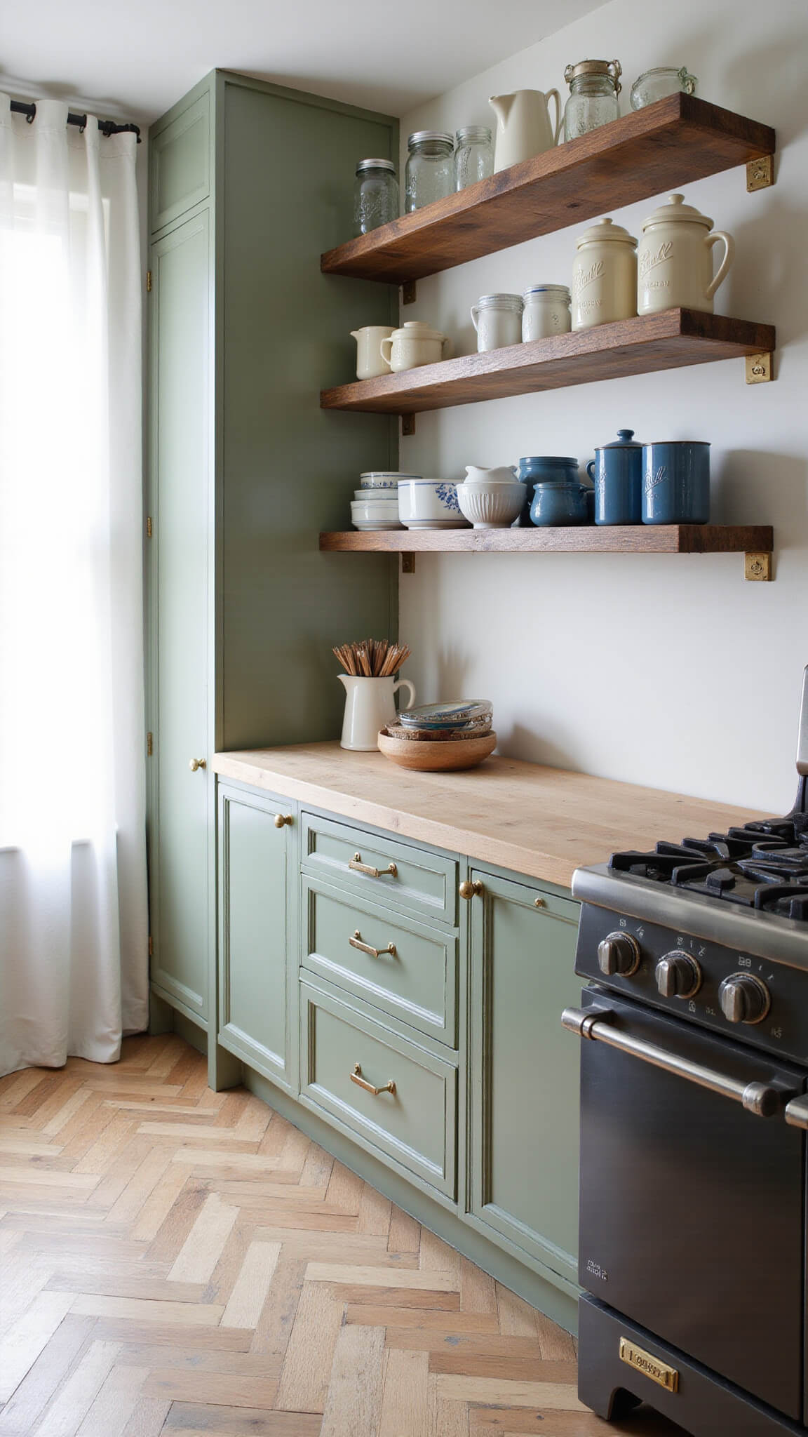 Low-angle shot of a rustic kitchen with sage green floor-to-ceiling cabinetry, brass pulls, reclaimed wood shelves holding ironstone pitchers and mason jars, worn butcher block countertops, vintage enamelware near a professional range, and herringbone wood flooring lit by morning sunlight through sheer curtains.