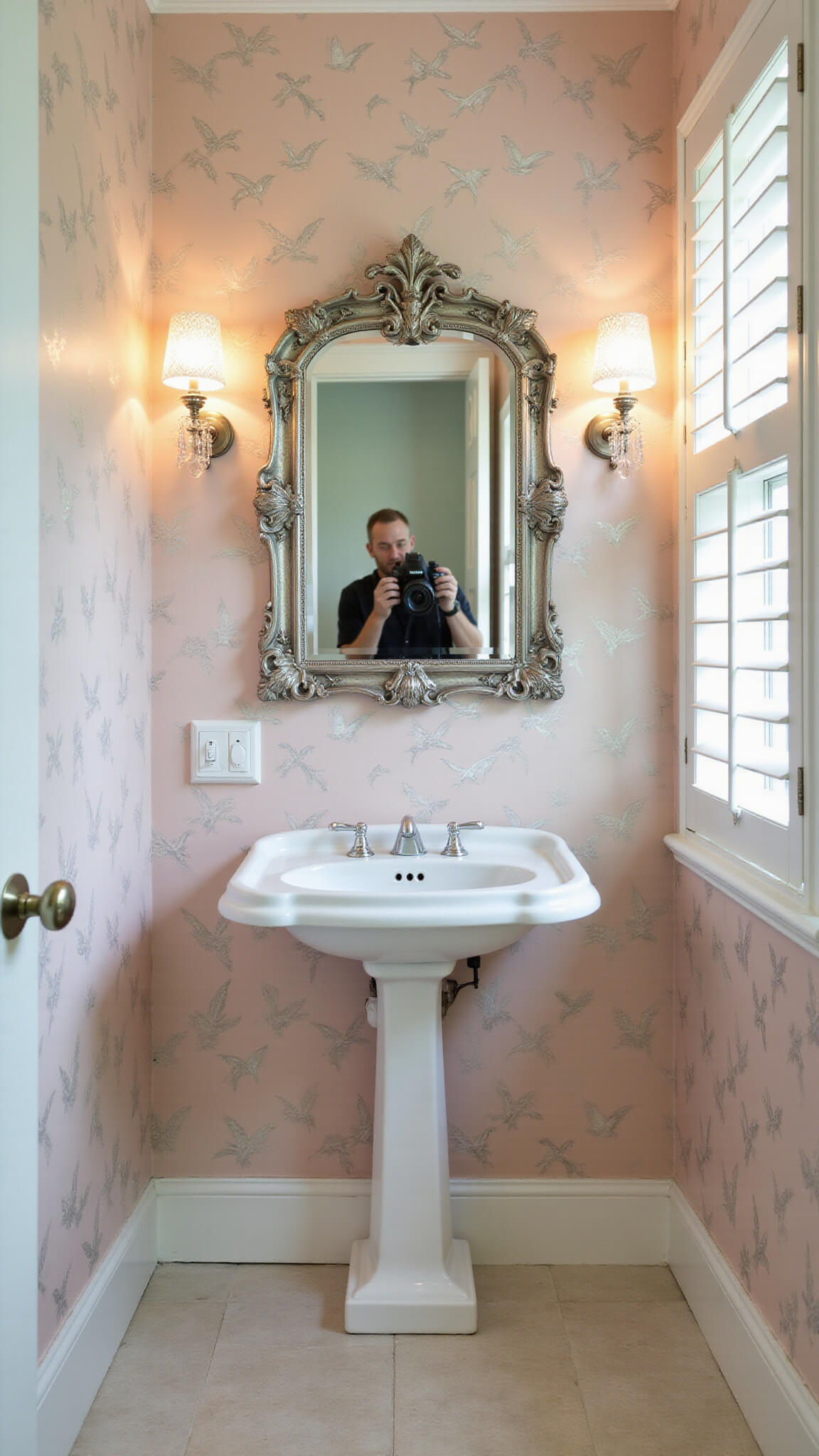 Small bathroom with blush and silver hummingbird wallpaper, white Victorian pedestal sink, ornate silver mirror, crystal sconces, and golden hour light through plantation shutters.