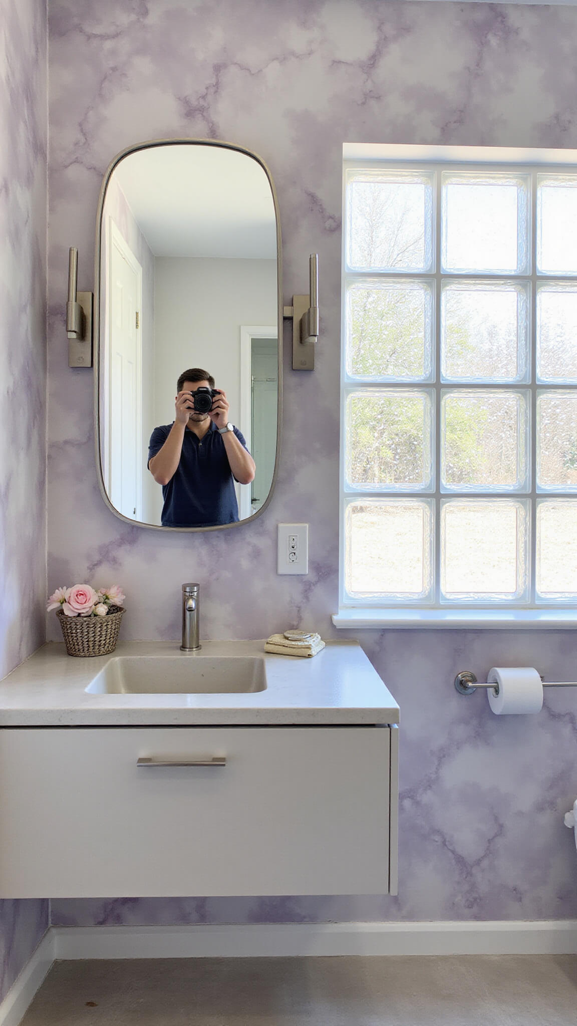 Modern 5x6ft bathroom with lavender and silver watercolor wallpaper, concrete wall-hung vanity, full-width mirror, and natural morning light through glass blocks.