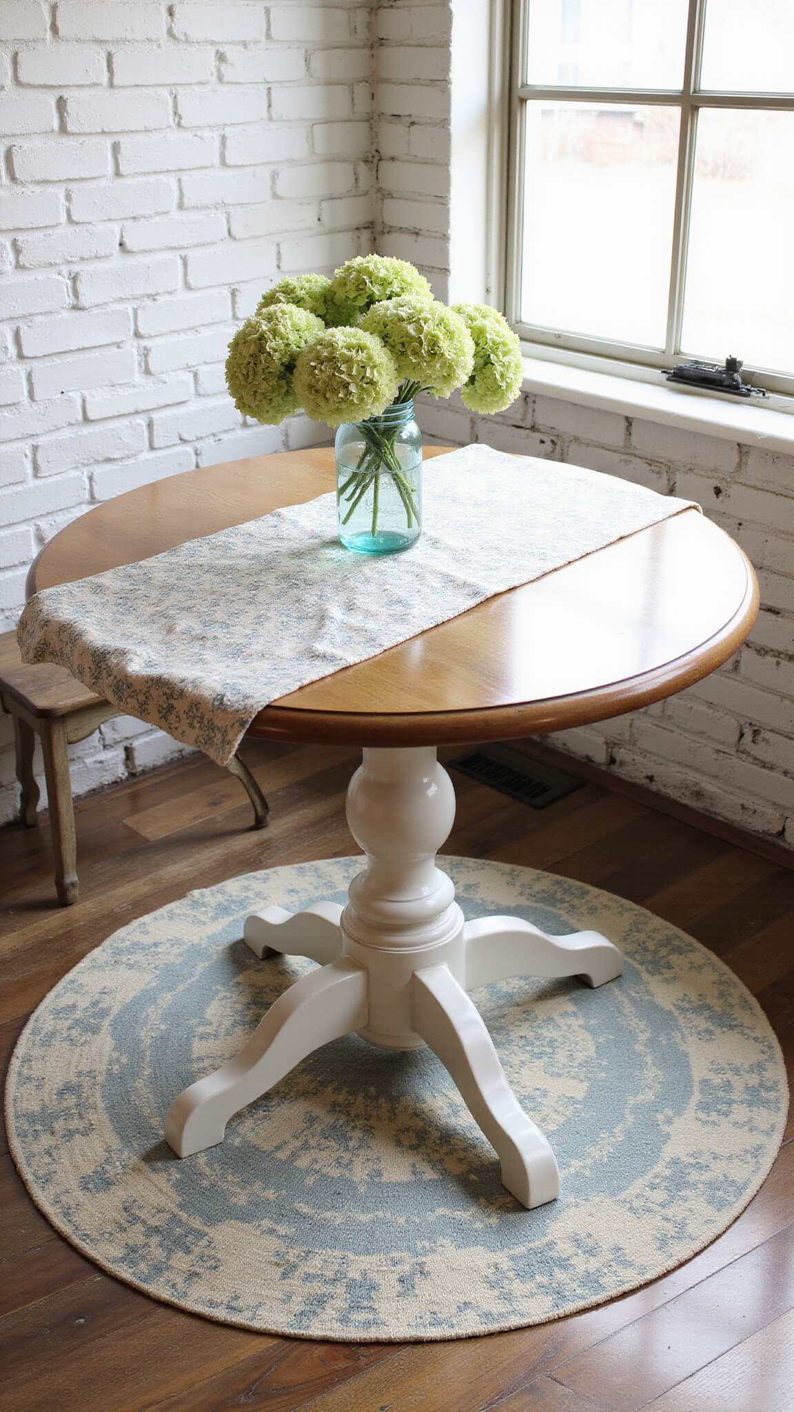 Overhead view of cozy kitchen nook with vintage floral farmhouse mat under rustic round table, whitewashed brick walls, reclaimed wood floor, and morning light casting shadows from hydrangeas in mason jar.