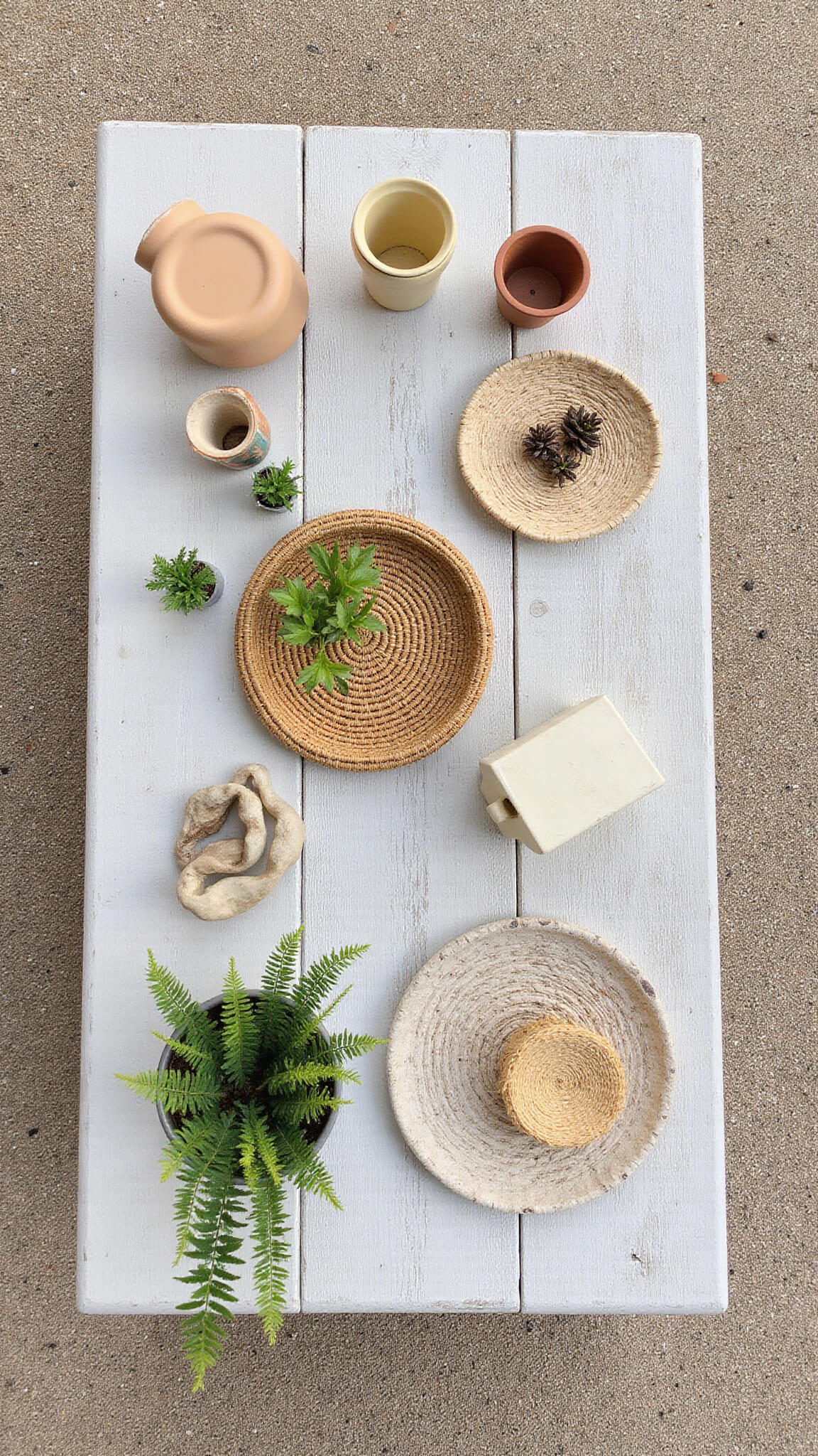 Flat lay of a styled patio vignette with woven baskets, ceramic planters, DIY painted pots, and small plants on a distressed wood table in bright, indirect light.