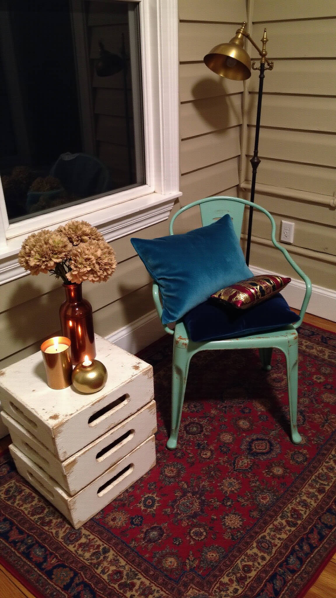 Moody dusk porch scene with turquoise metal chair, velvet pillows, Persian rug, brass lamp, and rustic decor.