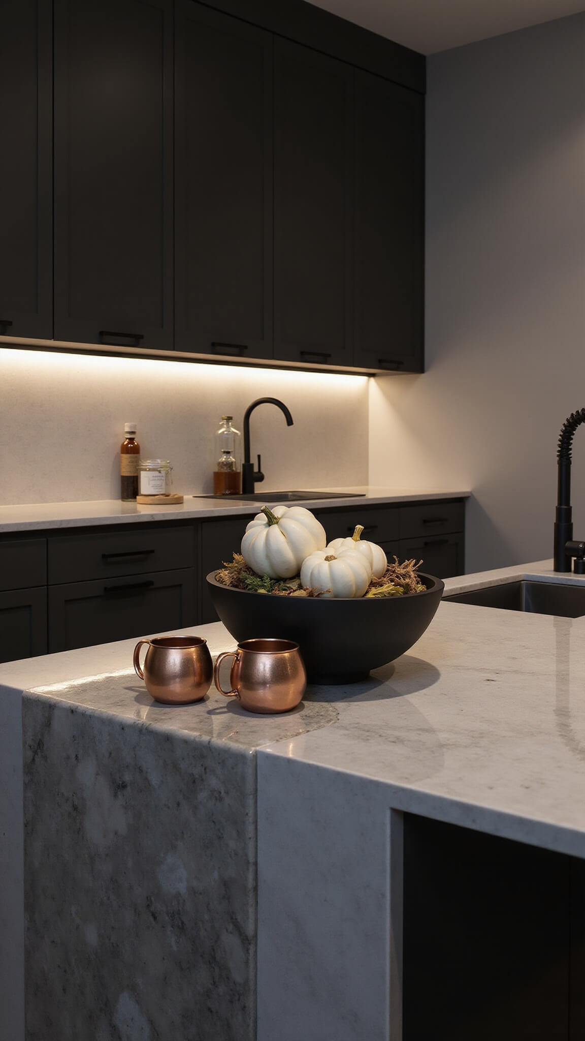 Modern kitchen at dusk with dark charcoal cabinets, marble countertops, warm LED lighting, and minimalist fall decor on a waterfall island.