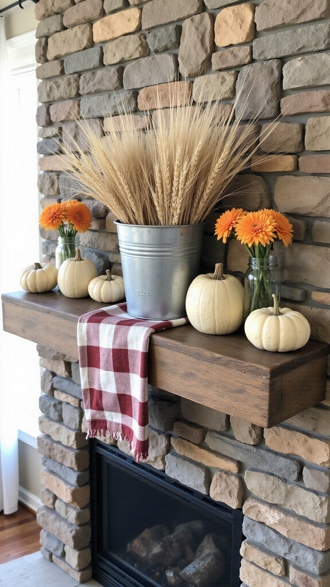 Rustic farmhouse fireplace with autumn decor, including wheat stalks in a metal container, cream pumpkins, orange dahlias in mason jars, and a buffalo check runner, lit by late afternoon sunlight.
