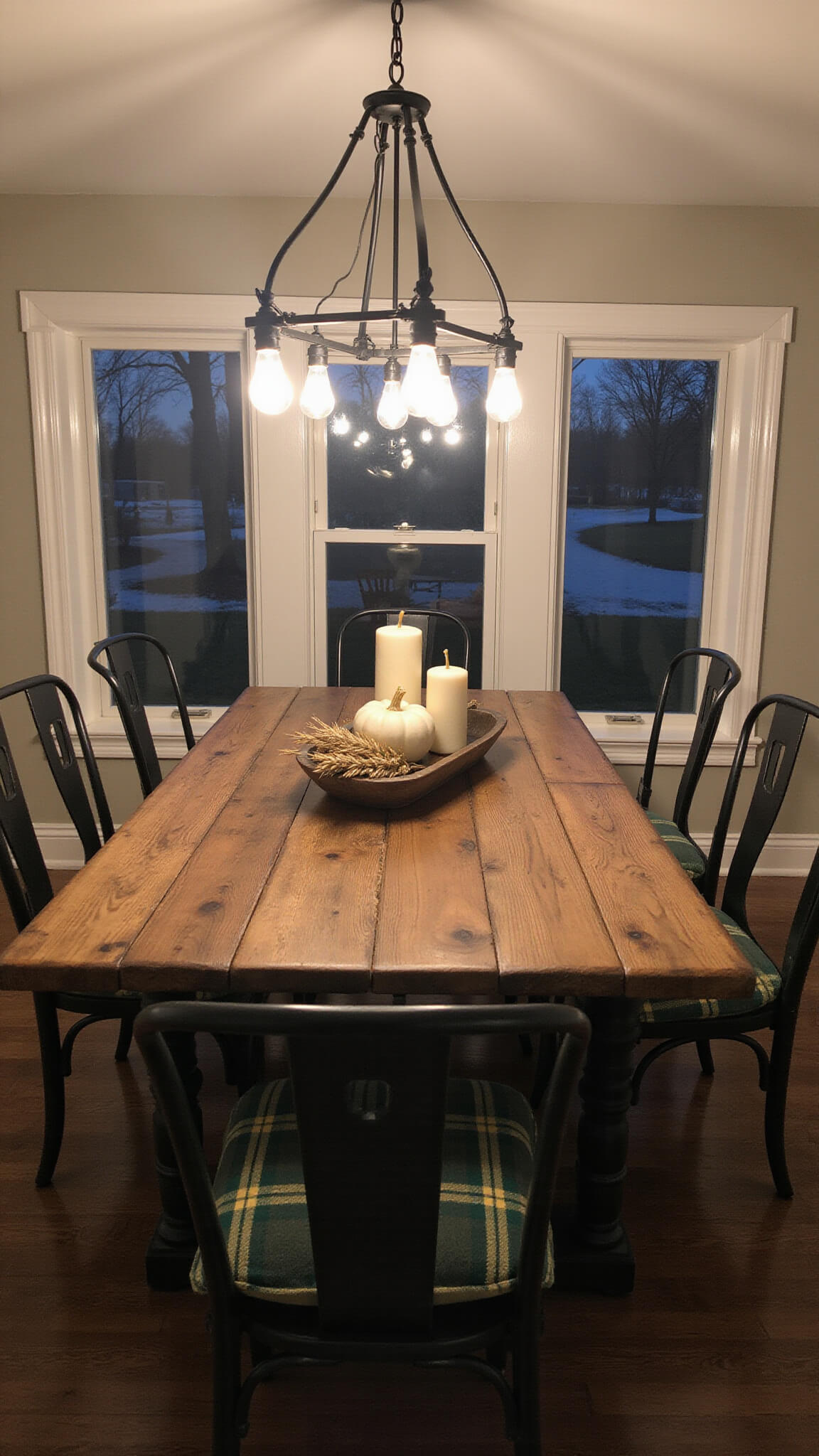 Modern farmhouse dining room at dusk with reclaimed wood table, white pumpkins, mercury glass candles, dried wheat centerpiece, plaid-cushioned metal chairs, and moody Edison bulb chandelier.