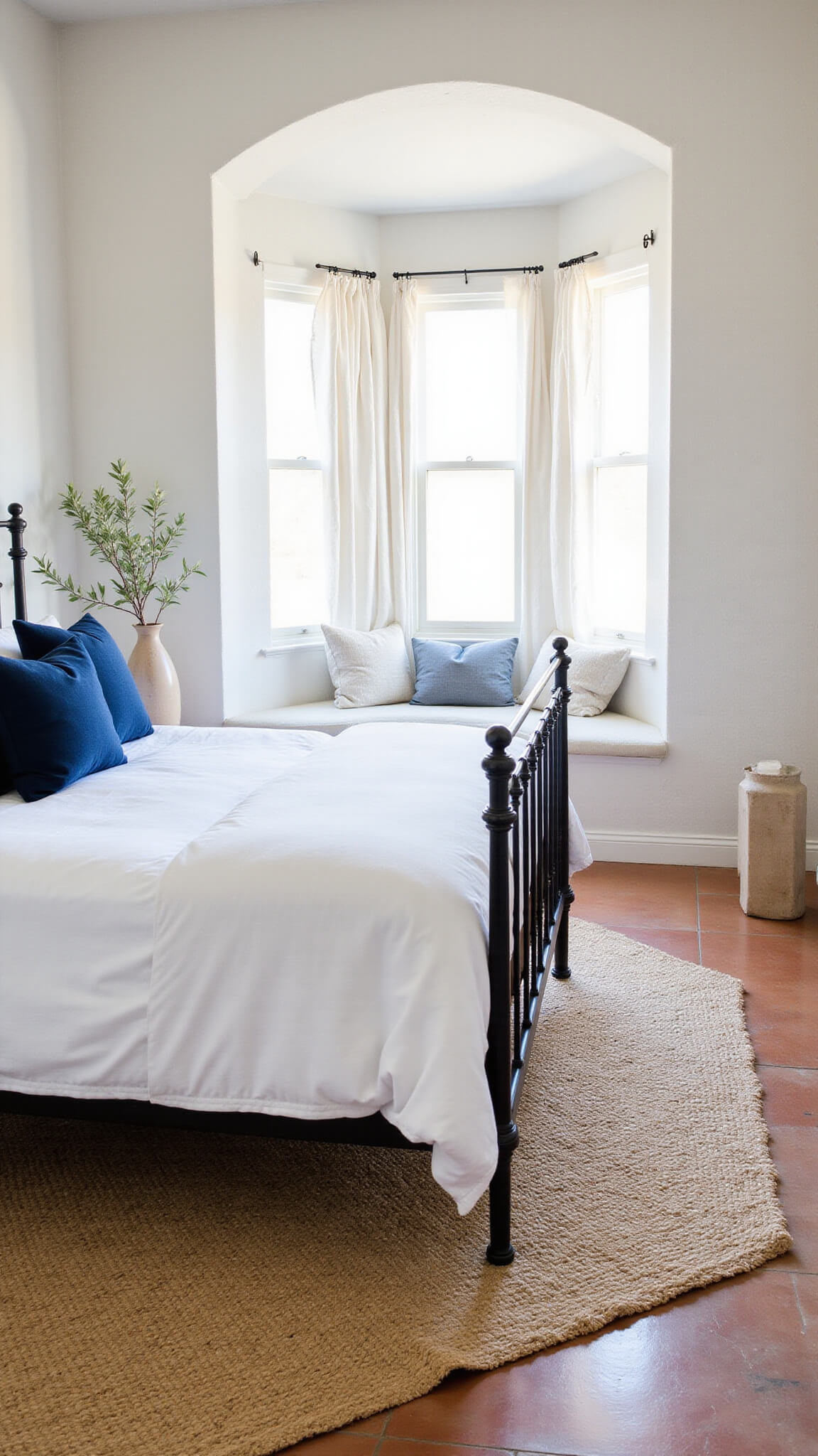 Airy Mediterranean bedroom with whitewashed textured walls, arched window alcove, wrought iron bed, terracotta floors, and morning light filtering through sheer white curtains.