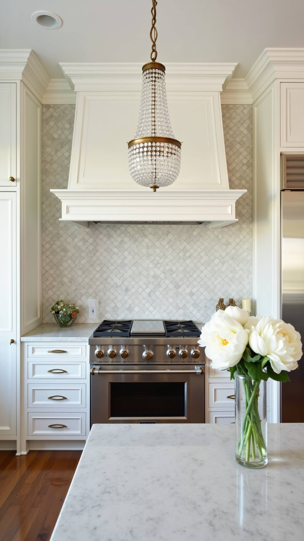 Elegant traditional kitchen with marble herringbone backsplash, crystal chandelier, white inset cabinets, brass accents, and white peonies in a mercury glass vase.