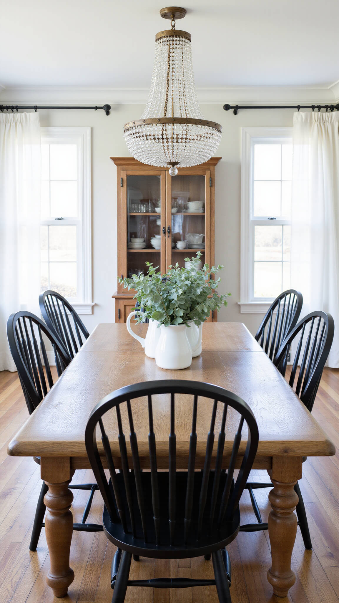 Elegant dining room with weathered oak farmhouse table, matte black Windsor chairs, eucalyptus-filled white pitchers centerpiece, and vintage crystal chandelier in soft mid-morning light.