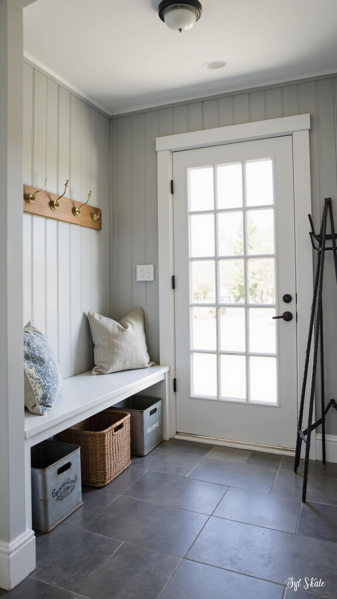 Functional mudroom with gray board and batten walls, slate flooring, built-in bench with brass hooks and vintage crates, woven baskets, and black iron coat rack in warm afternoon light.