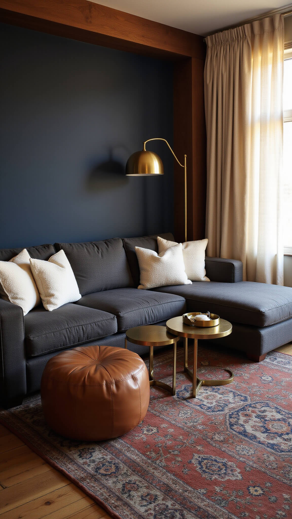 Cozy 16x14ft lounge at sunset with charcoal sectional, ivory bouclé pillows, navy accent wall, brass floor lamp, layered vintage rugs, walnut media unit, cream curtains, leather poufs, and brass side tables.