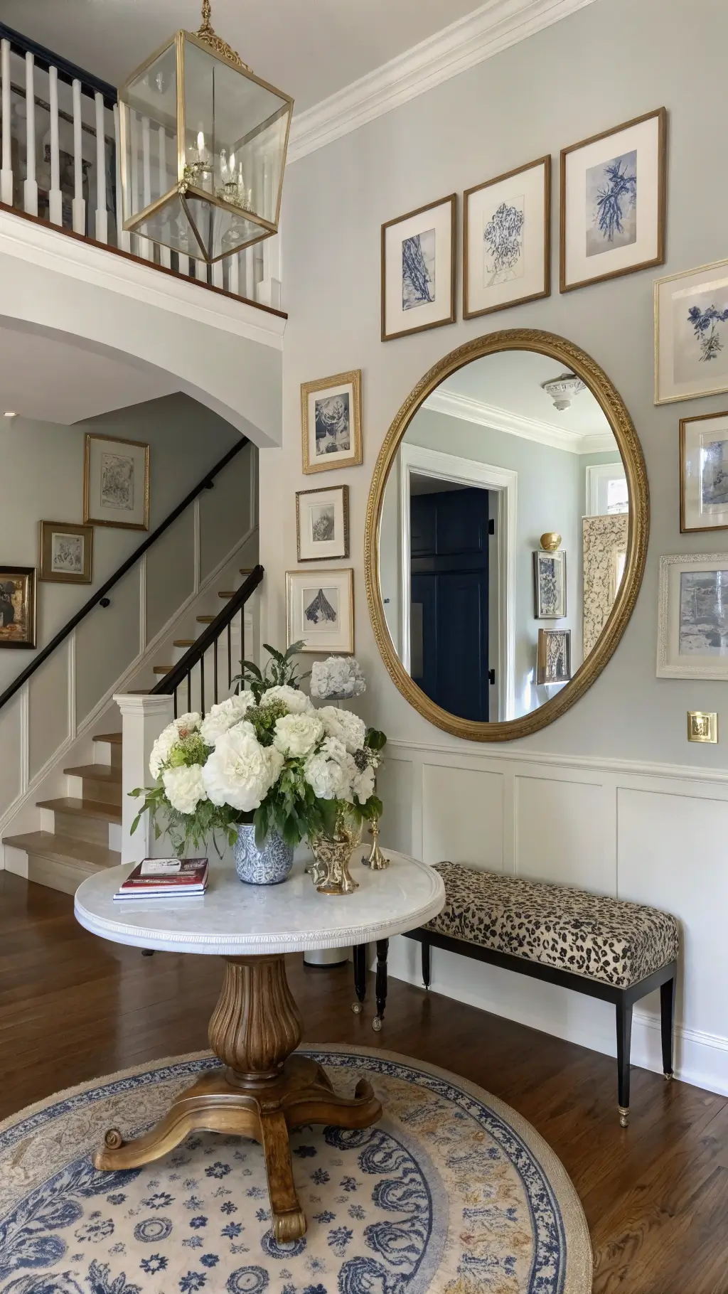 Elegant entryway with round table holding large floral arrangement, oversized mirror, blue and white chinoiserie, gallery wall on staircase, vintage runner, brass umbrella stand, leopard print bench, and morning light illuminating navy, white, and brass accents.