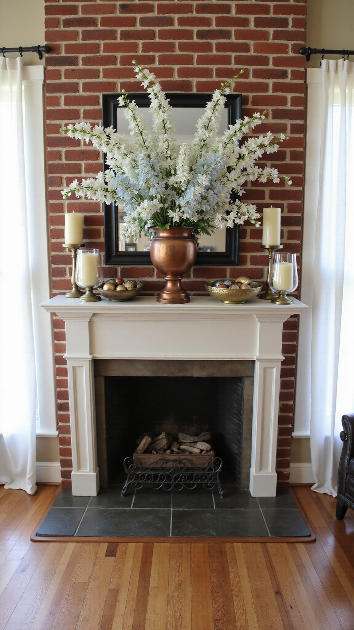 Close-up of spring-themed fireplace mantel with white cherry blossoms, blue delphiniums in a copper urn, metallic eggs in brass bowls, and ivory candles in glass hurricanes illuminated by soft morning light.