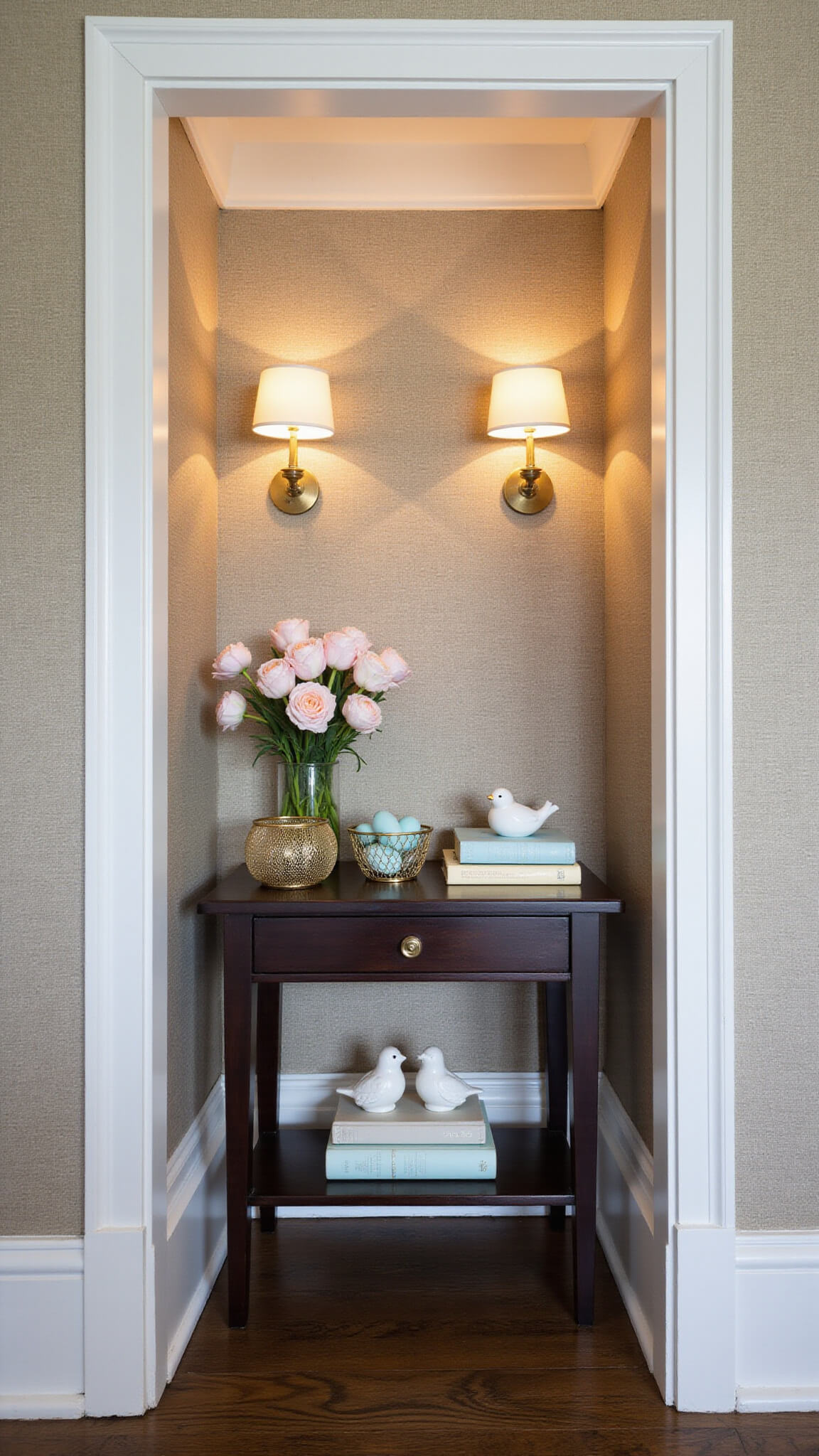 Entry alcove with grasscloth walls, warm sconce lighting, dark wood console table displaying pale flowers, gold baskets with blue eggs, and ceramic birds on vintage books.