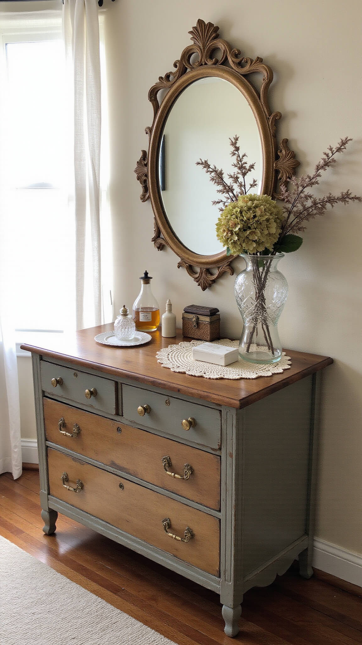 Sunlit vintage bedroom nook with a weathered pine dresser, oval gilded mirror, crystal trinkets, dried hydrangeas, and antique brass details.