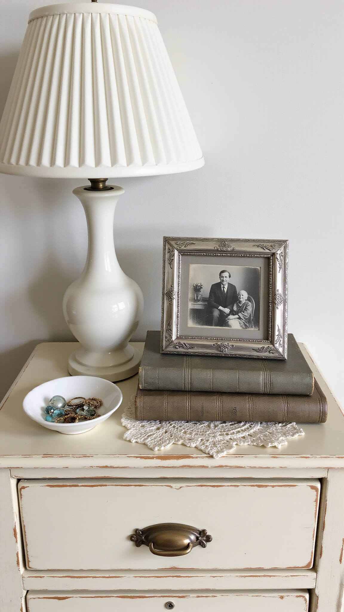 Overhead shot of a vintage cream nightstand at 9am featuring a milk glass lamp, antique silver photo frame, worn hardcover books, porcelain jewelry dish, and hand-tatted doily in soft morning light with shallow depth of field.