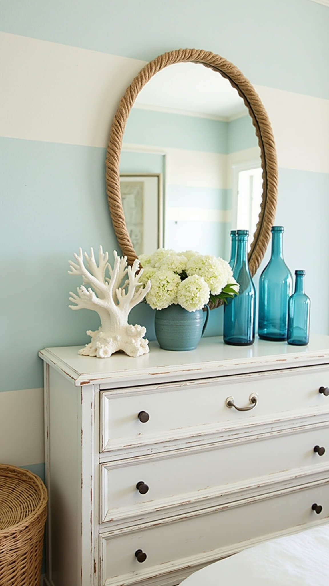 Coastal bedroom dresser vignette with round rope-framed mirror, white coral, blue glass bottles, white hydrangeas, and textured basket in soft morning light.