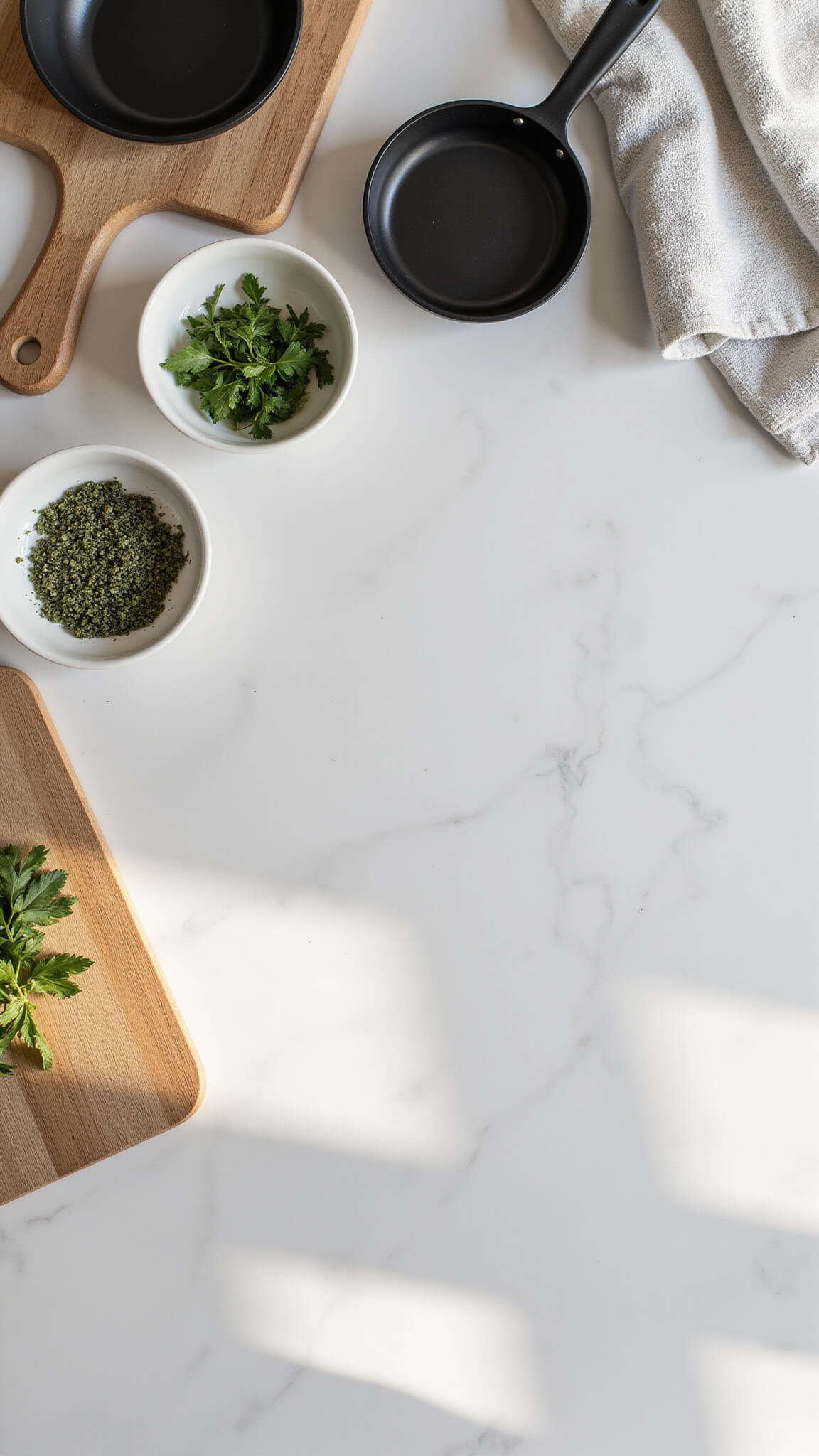 Overhead view of a Nordic kitchen workspace with white marble counter, wooden cutting boards, matte black cookware, linen tea towels, and three ceramic bowls of fresh herbs, lit by soft morning light.