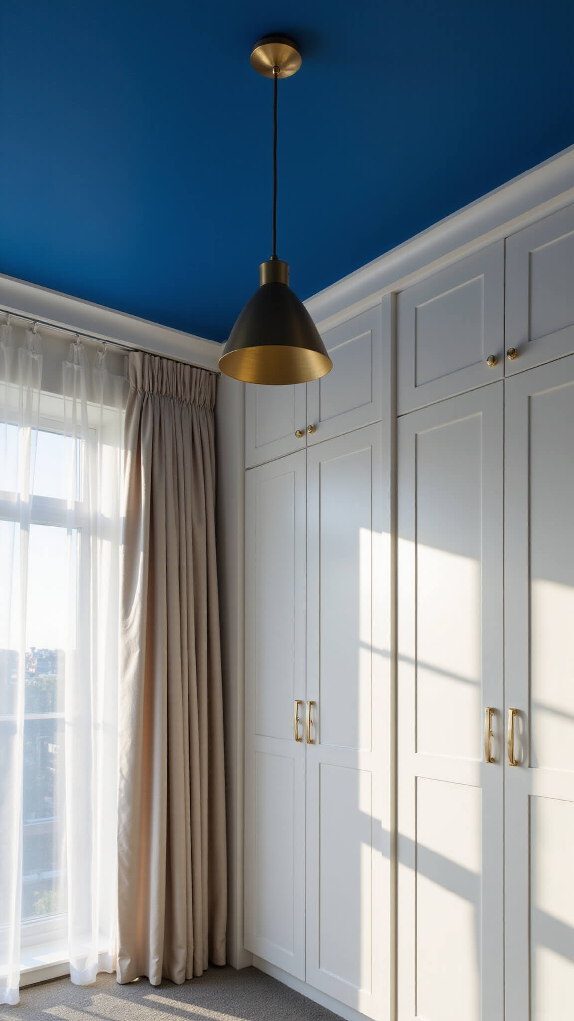 Wide-angle view of white bedroom with 10ft sapphire blue ceiling, brushed gold pendant light, built-in wardrobes, and dramatic sunlight through sheer curtains.