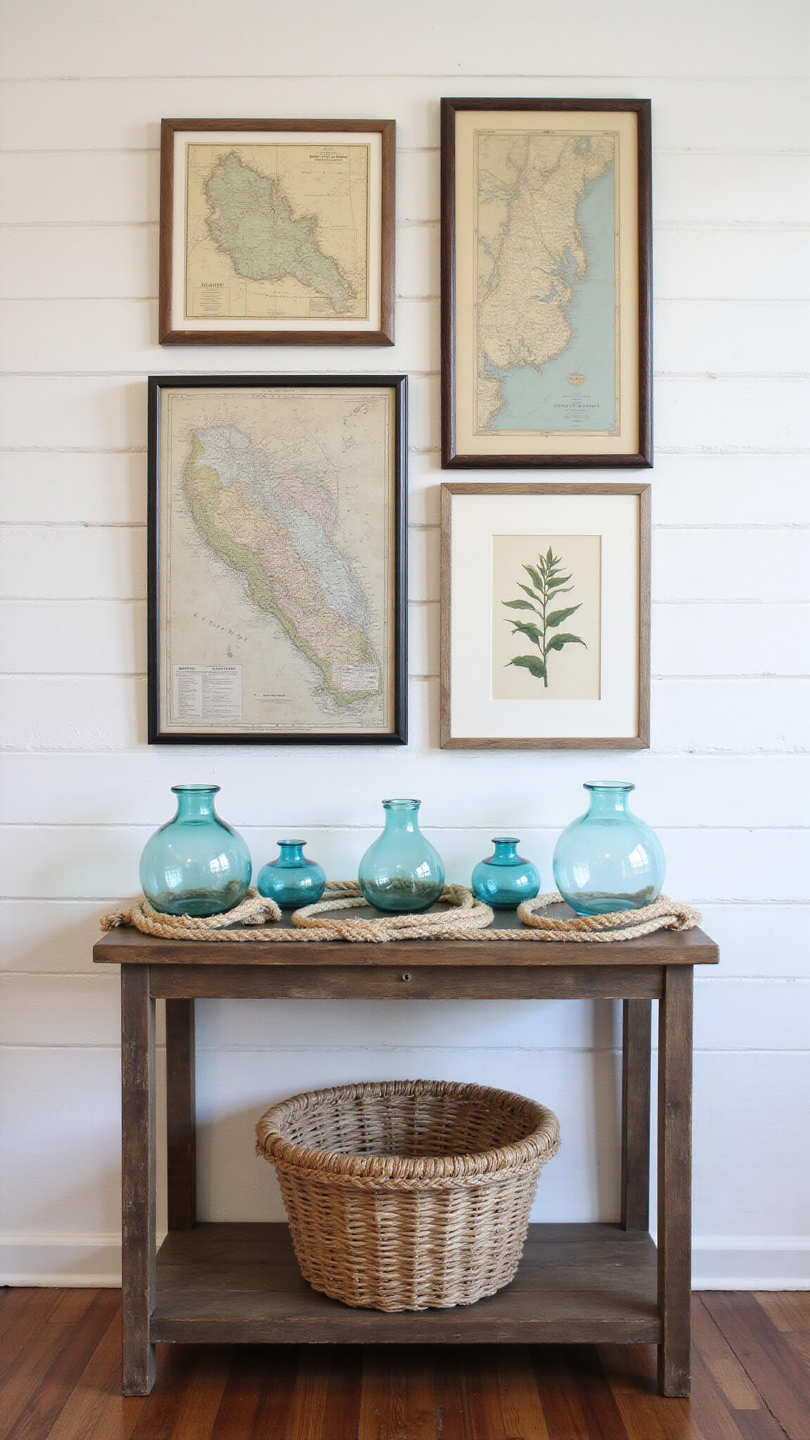 Entryway gallery wall with vintage naval maps and botanical prints in distressed frames on whitewashed shiplap; reclaimed wood console table with blue glass vessels, rope knots, and seagrass basket underneath.